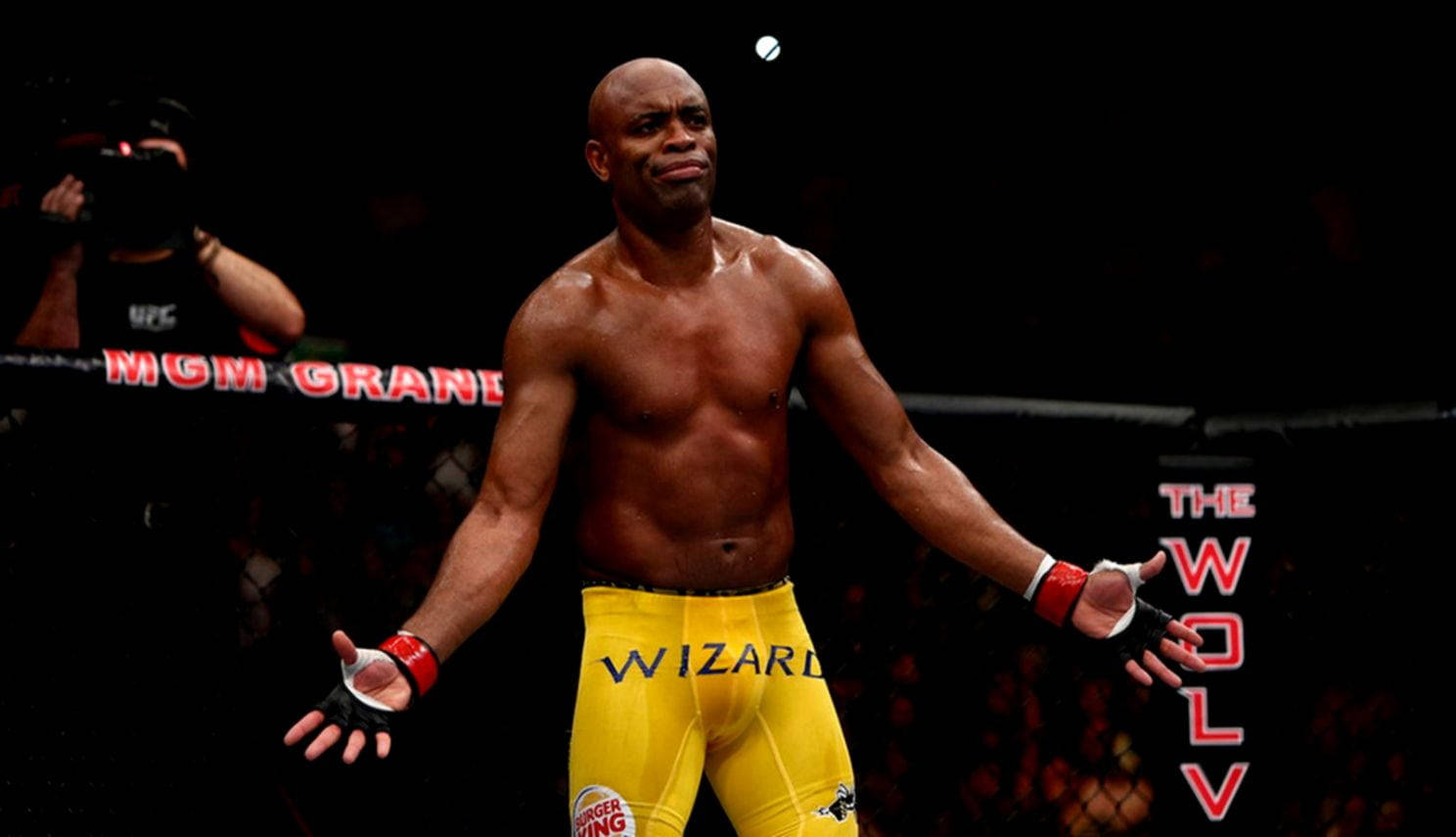 一位勝利的MMA拳手穿著黃色短褲，在八角籠中張開雙臂站立。