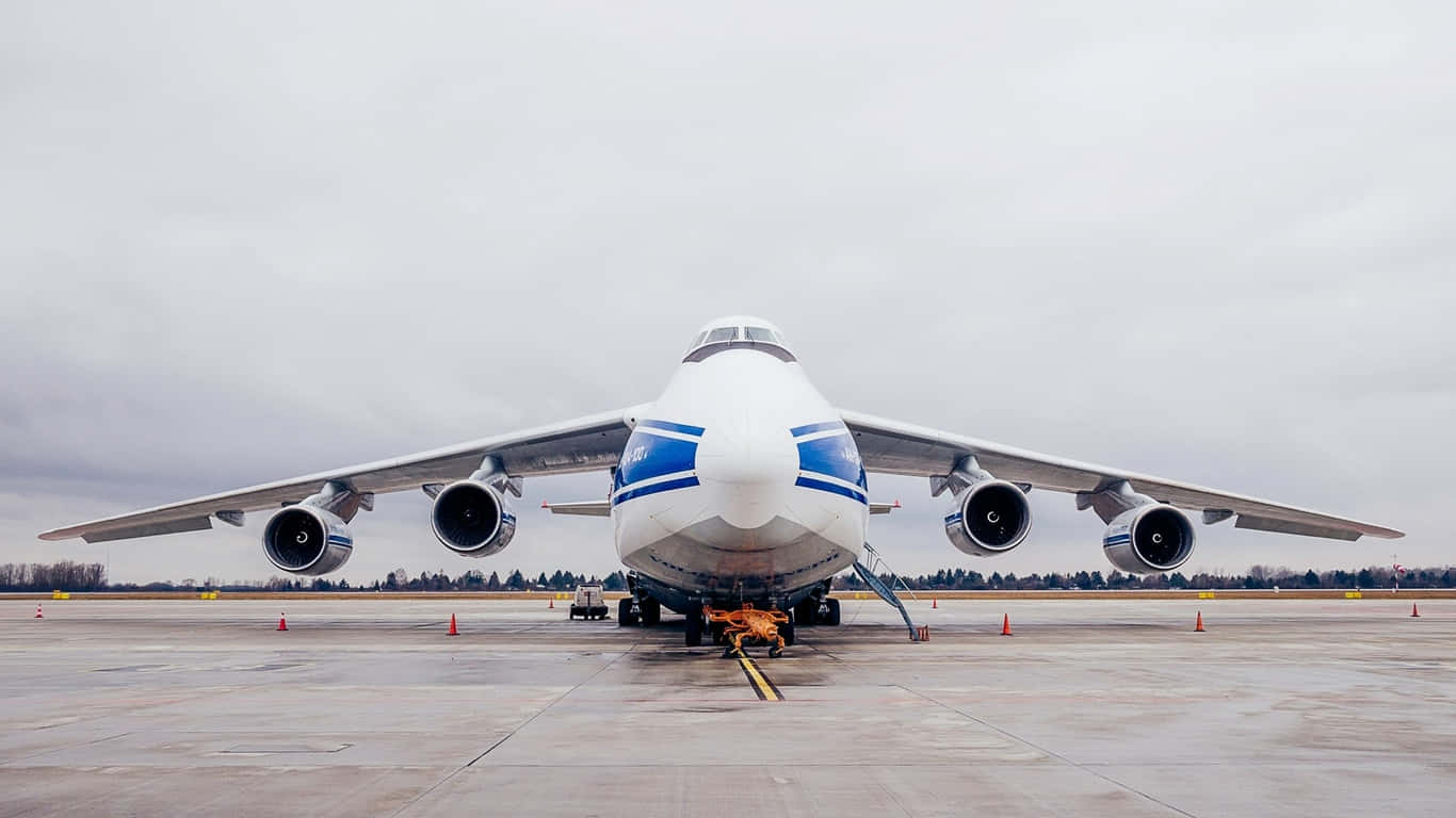 安托諾夫 An-124 羅斯蘭正面視角停泊於暴風雲背景的巨型客機背景（1366x768） 壁紙 — 一架巨大的白色和藍色貨機停在潮濕的停機坪上，天空多雲，附近有橙色的地勤設備。