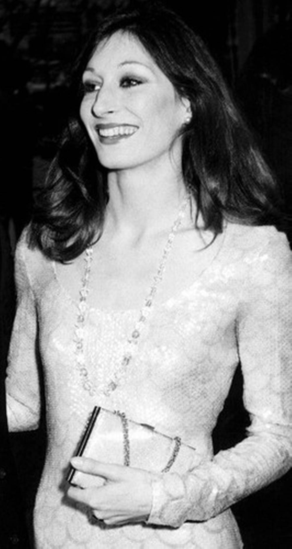 安潔莉卡·休斯頓 1974 年奧斯卡頒獎典禮 壁紙 — 一位微笑的女性穿著質感連衣裙和珍珠項鍊，在昏暗的環境中拿著手拿包。