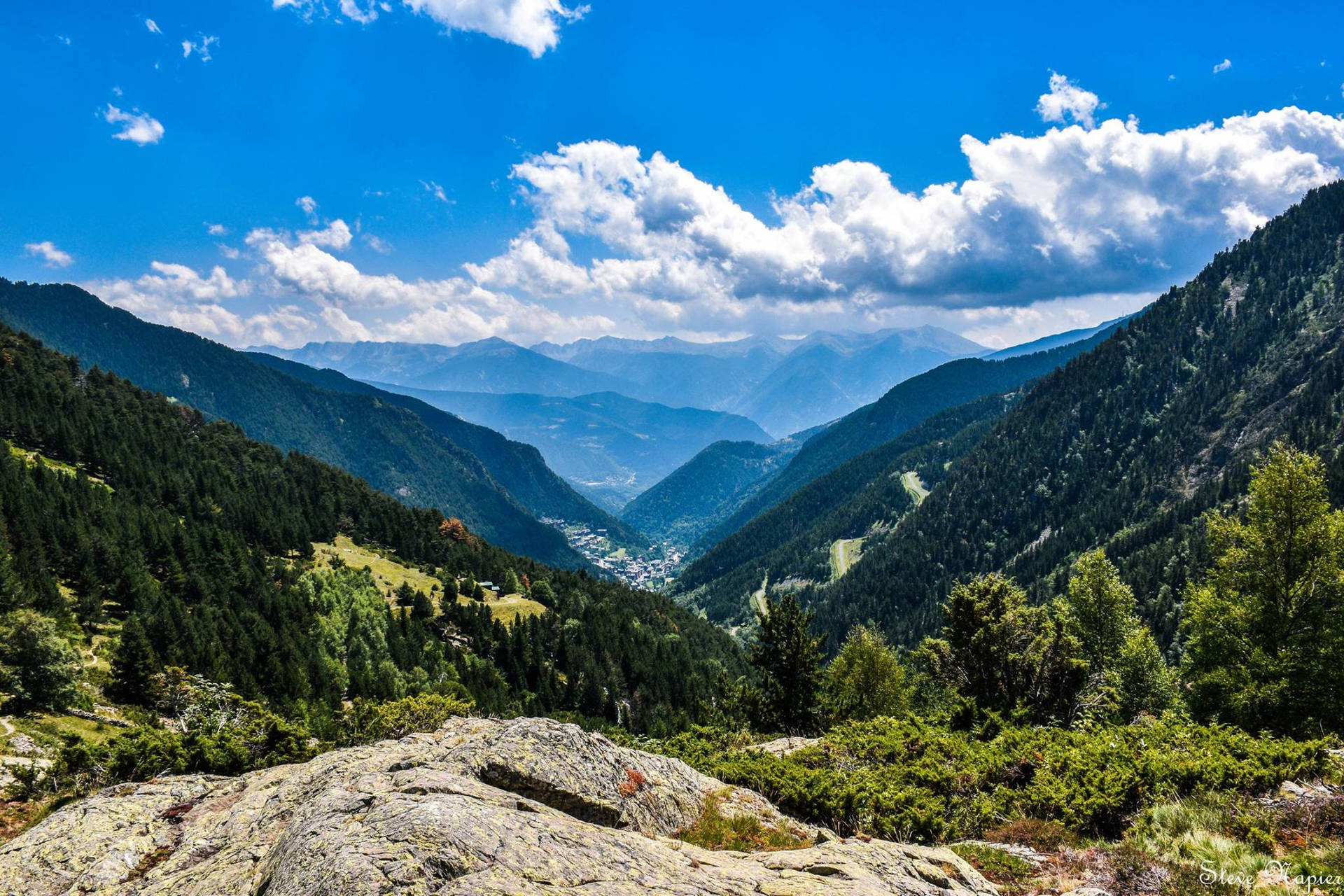 山谷的全景，森林覆蓋的山坡和下方的小鎮，在晴朗的藍天下。