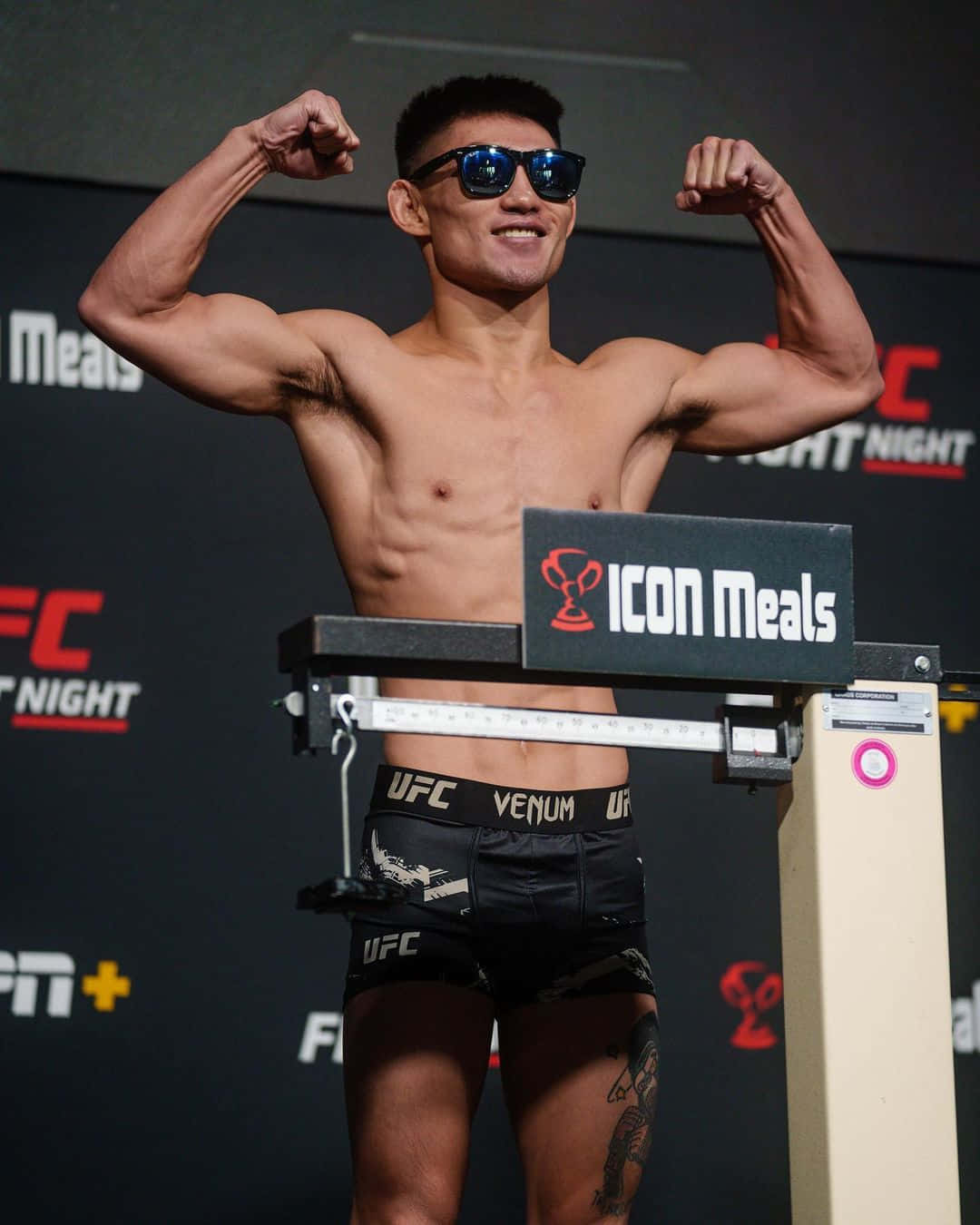 UFC拳手在稱重活動中站在體重秤上展示肌肉，戴著太陽眼鏡和品牌短褲。