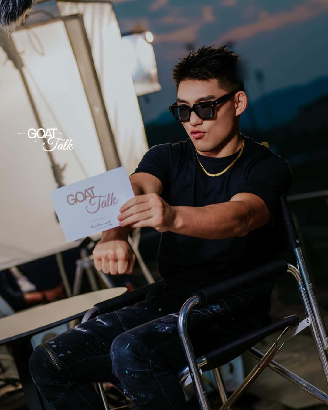 戴著太陽眼鏡的男士在導演椅上手持GOAT Talk卡片，於黃昏時分。