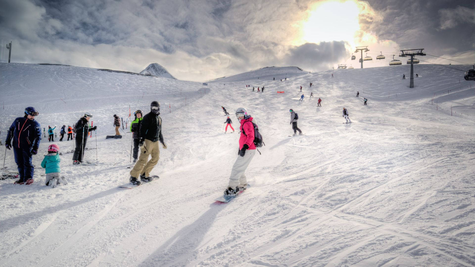 完美冬季坡地 壁紙 — 滑雪者和單板滑雪者在陽光普照的雪坡上滑行，天空有戲劇性的雲層，遠處有纜車。
