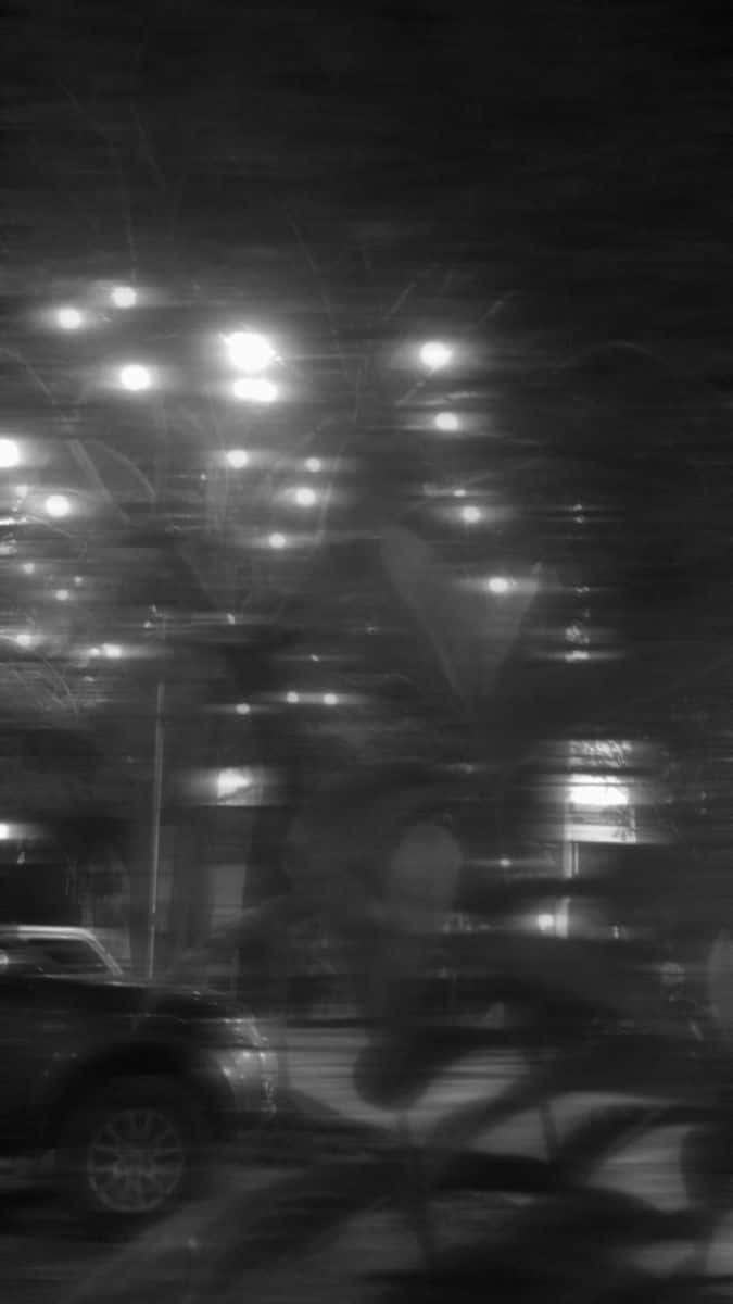雨痕模糊了夜間擋風玻璃上的城市燈光，可見一輛停著的車。