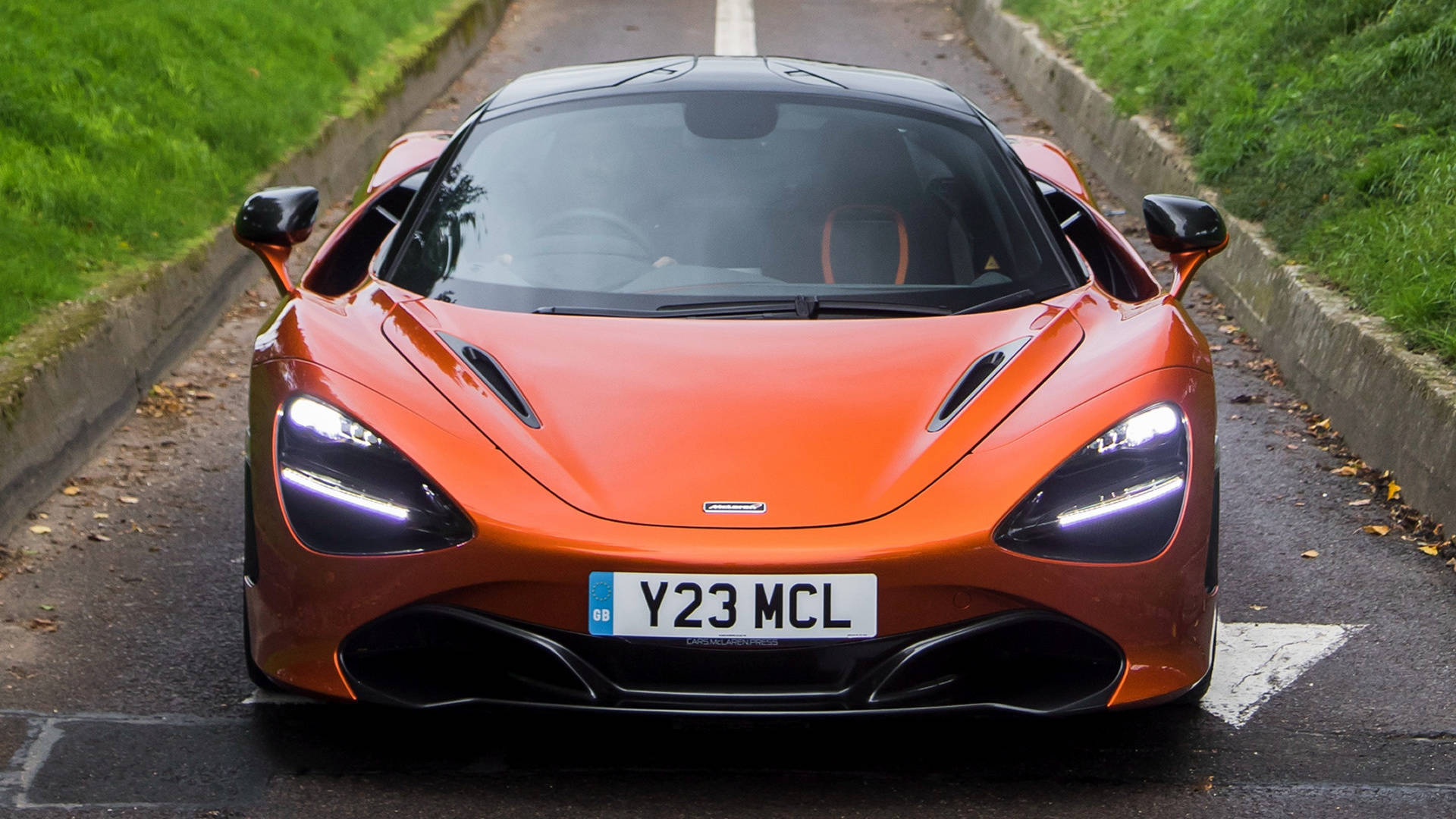 完美設計 - 麥拉倫720S 4K前視圖 壁紙 — 一輛充滿活力的橙色McLaren 720S跑車在濕滑的道路上向前行駛，兩旁是綠草和落葉。