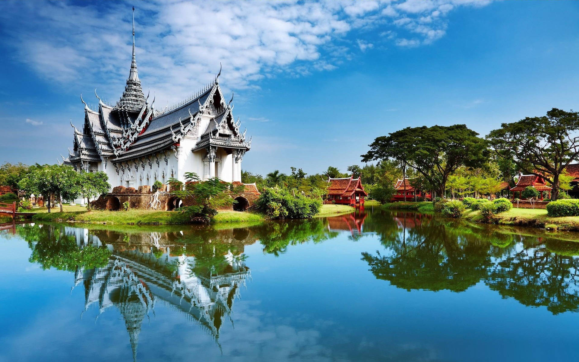 白色泰國寺廟，尖頂屋頂倒映在寧靜的池塘中，周圍環繞著樹木。
