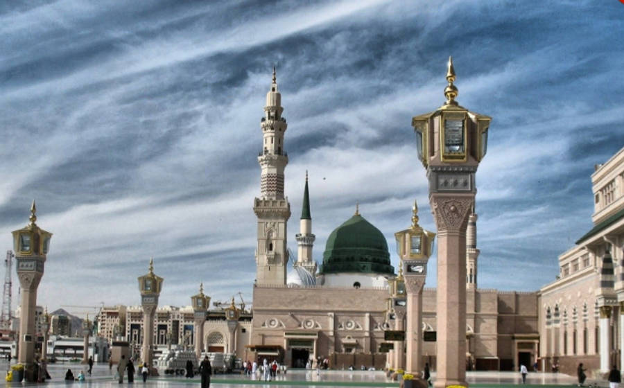 Madina 桌布 壁紙 — 宏偉清真寺，綠色圓頂和宣禮塔，戲劇性的多雲天空下，人們在庭院中行走。