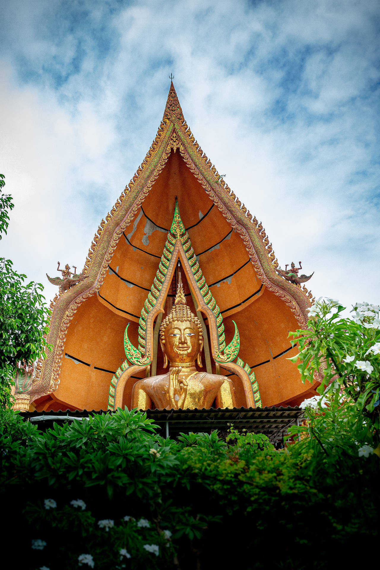 金色佛像在裝飾寺廟屋頂下，被綠色植物和雲朵環繞。
