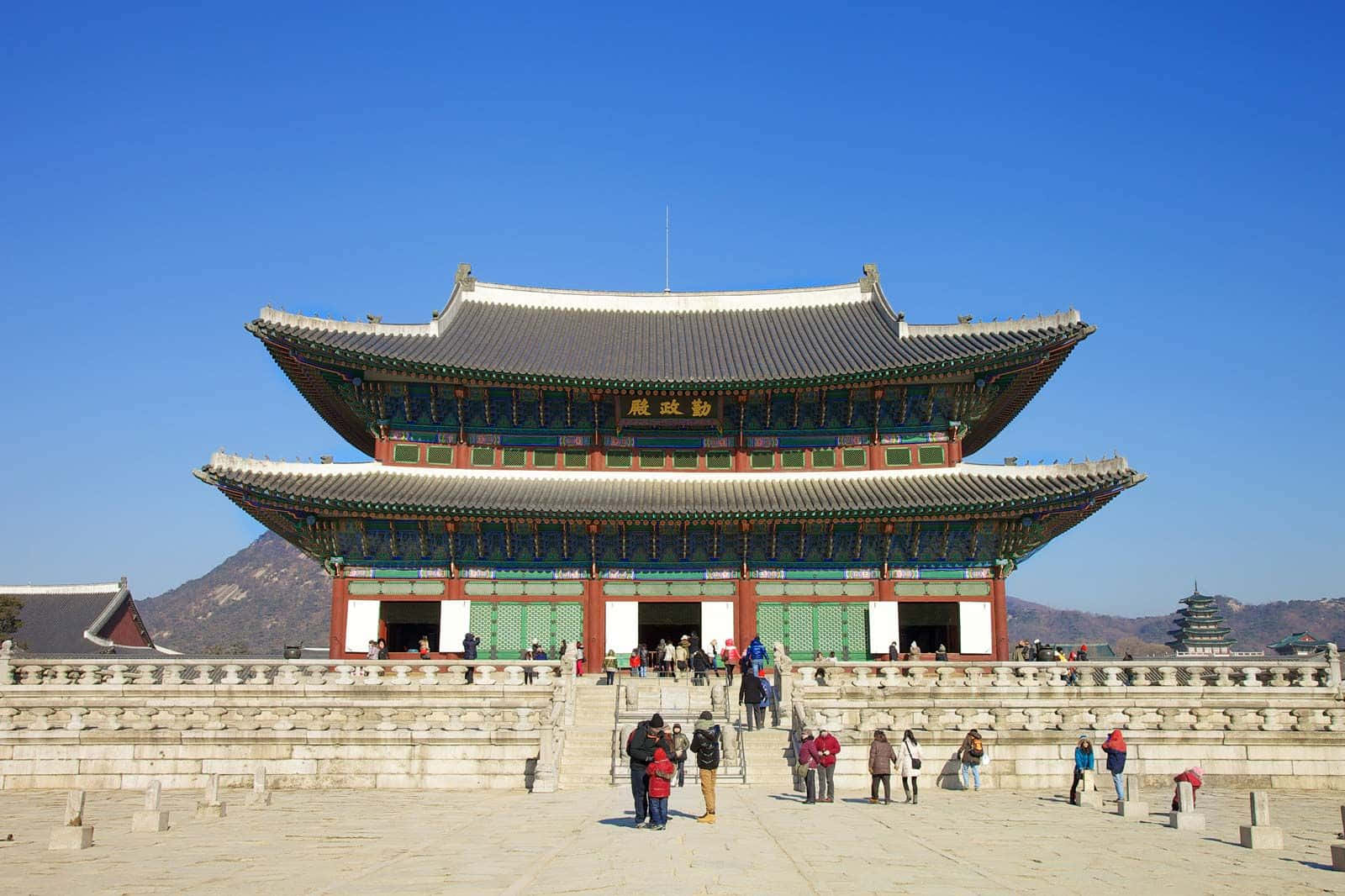 South Korea 桌布 壁紙 — 宏偉的傳統韓國宮殿，擁有蜿蜒的瓦片屋頂和紅色柱子，在清澈的藍天下，遊客正在探索石頭庭院。