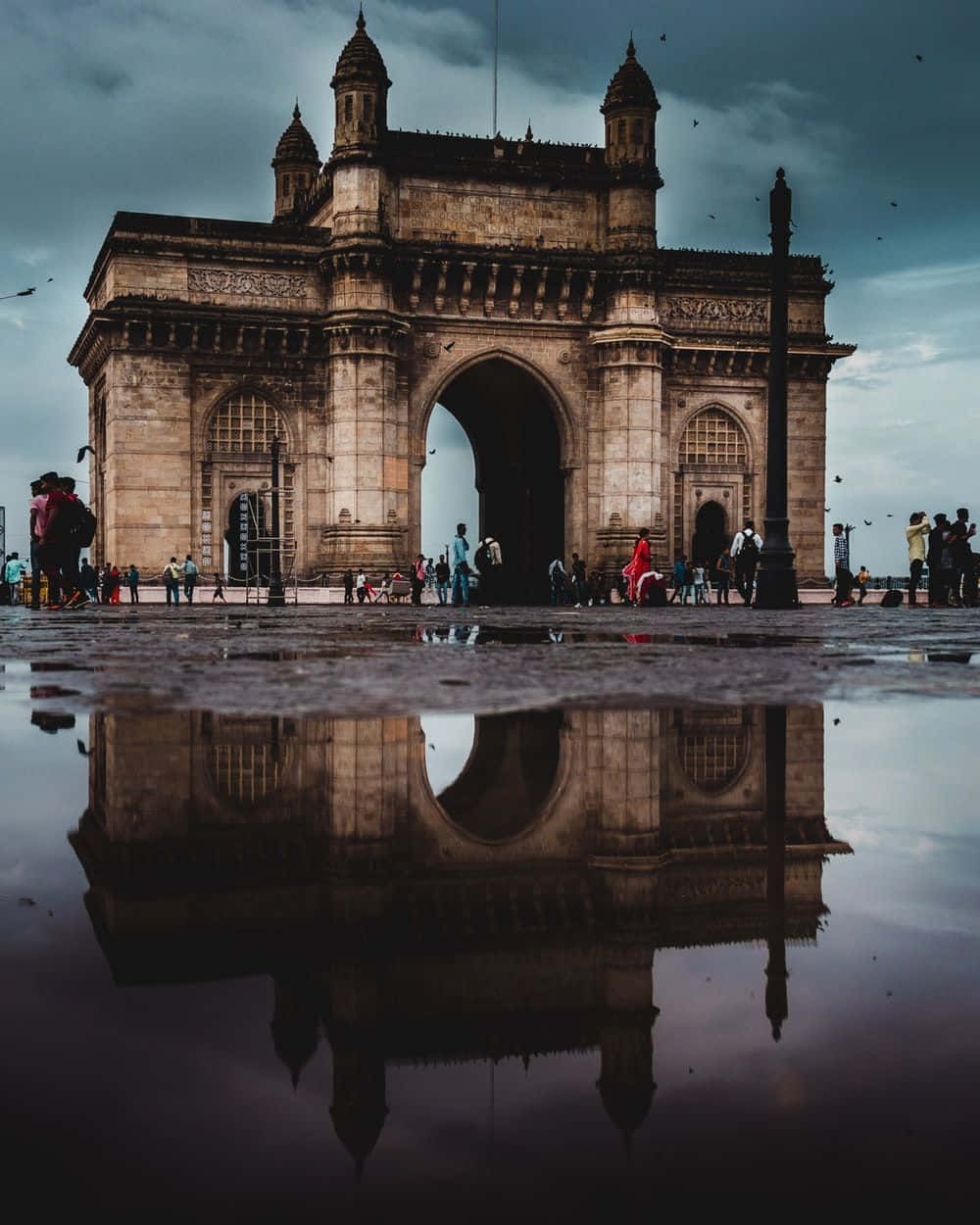 Mumbai 桌布 壁紙 — 宏偉的印度門紀念碑倒映在雨後水窪中，天空陰沉，附近有遊客。