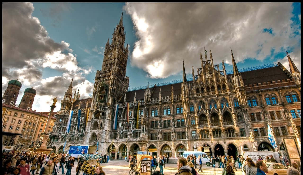Germany 桌布 壁紙 — 宏偉的哥德式市政廳，擁有尖頂和拱形窗戶，在戲劇性的多雲天空下主宰著繁忙的廣場。