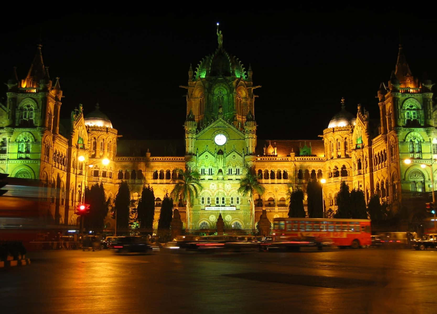 Mumbai 桌布 壁紙 — 宏偉的歷史火車站在夜晚以綠色和金色燈光照明，模糊的車流疾馳而過。