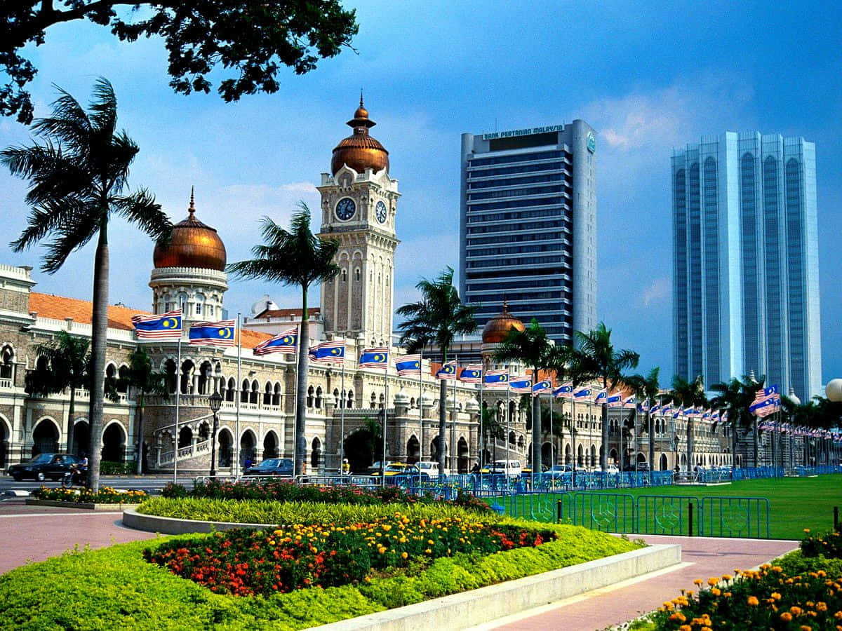 Malaysia 桌布 壁紙 — 宏偉的殖民風格鐘樓，金色圓頂，矗立在現代摩天大樓旁，藍天白雲，棕櫚樹和色彩繽紛的花壇。
