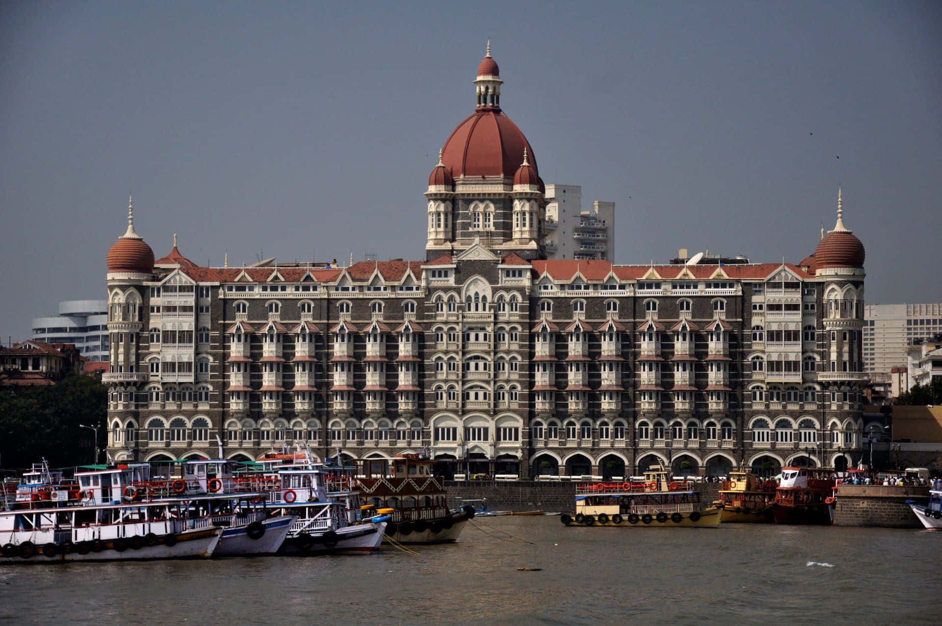 Mumbai 桌布 壁紙 — 宏偉的泰姬瑪哈皇宮酒店，紅色圓頂雄偉地矗立在海濱，船隻停泊在平靜的河邊，天空朦朧。