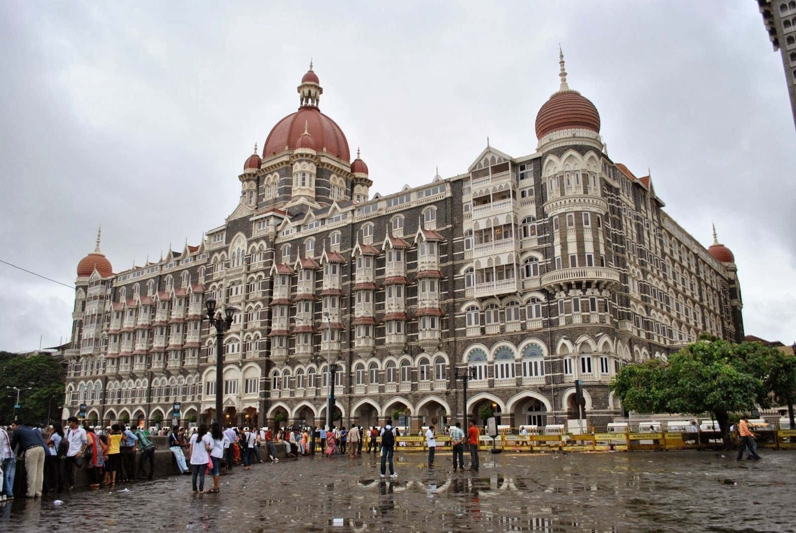 Mumbai 桌布 壁紙 — 宏偉的泰姬瑪哈皇宮酒店，紅色圓頂和華麗建築矗立在陰天之下，人們聚集在濕漉漉的路面上。