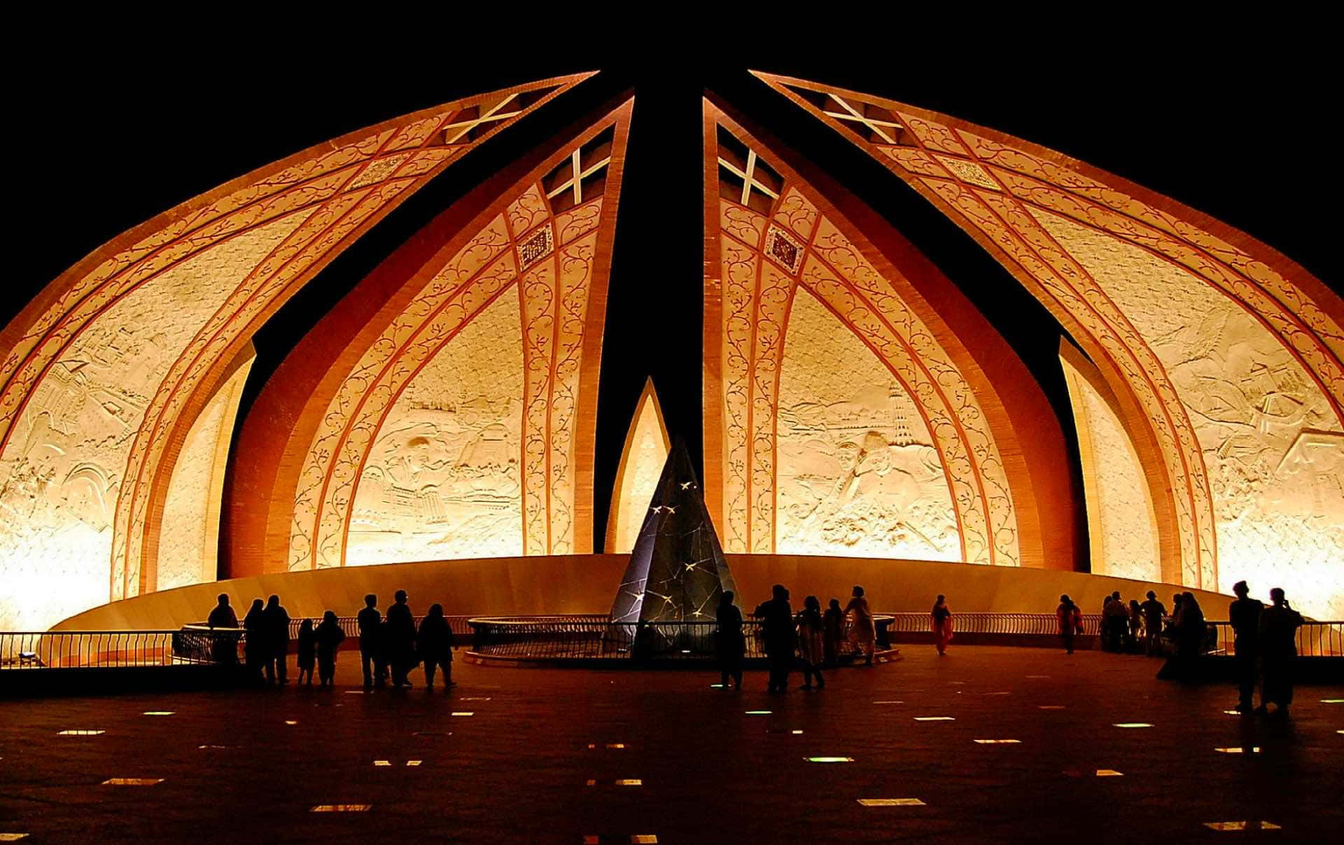 Pakistan 桌布 壁紙 — 宏偉的燈光清真寺，帶有發光的橙色拱門和複雜的雕刻，在夜晚莊嚴聳立，剪影般的遊客聚集在其基部周圍。