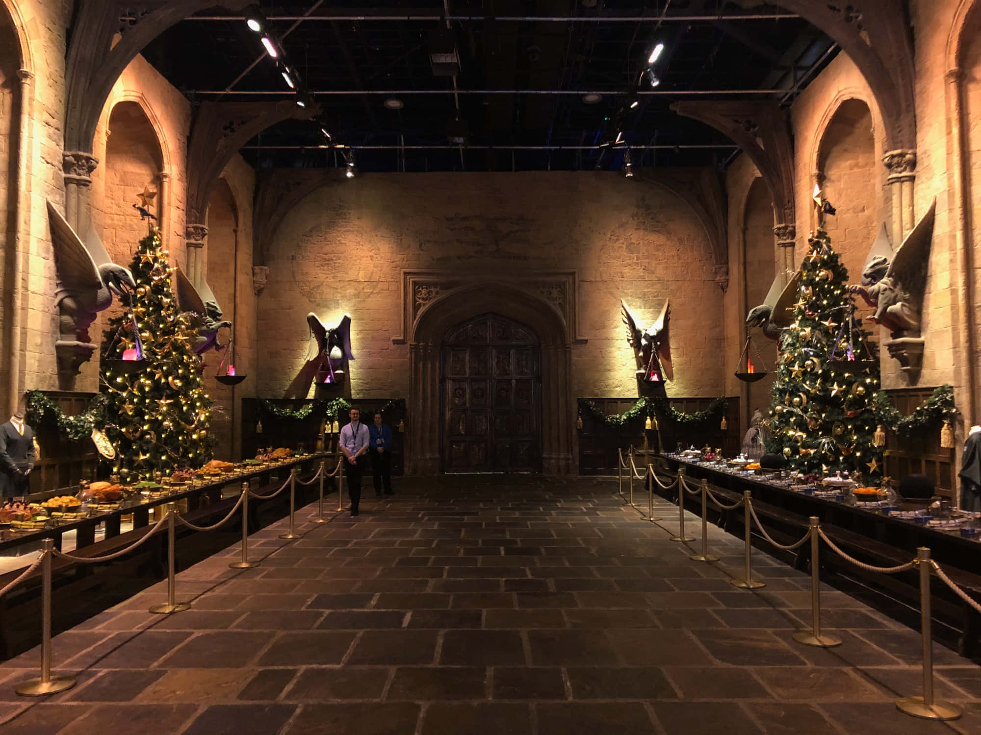 Harry Potter Christmas 桌布 壁紙 — 宏偉的石頭大廳，高聳的拱門和節日聖誕樹在溫暖的聚光燈下排列在長長的宴會桌兩旁。