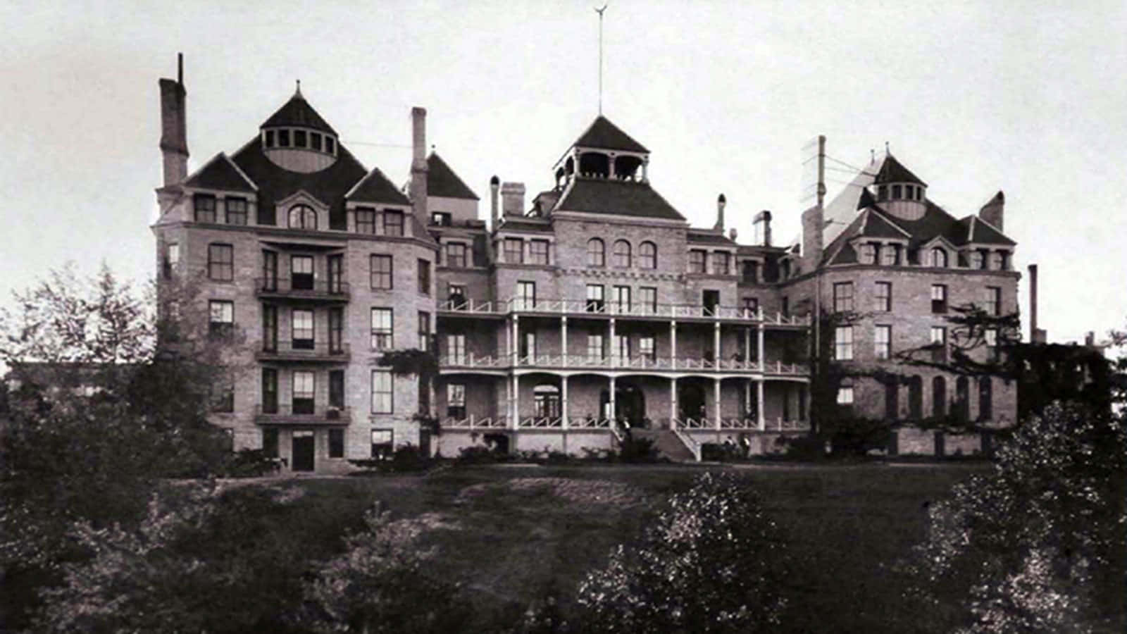 Haunted Hotels 桌布 壁紙 — 宏偉的維多利亞式大宅，設有塔樓和環繞式門廊，在泛黃的歷史照片中，屹立於樹木之間，氣勢非凡。