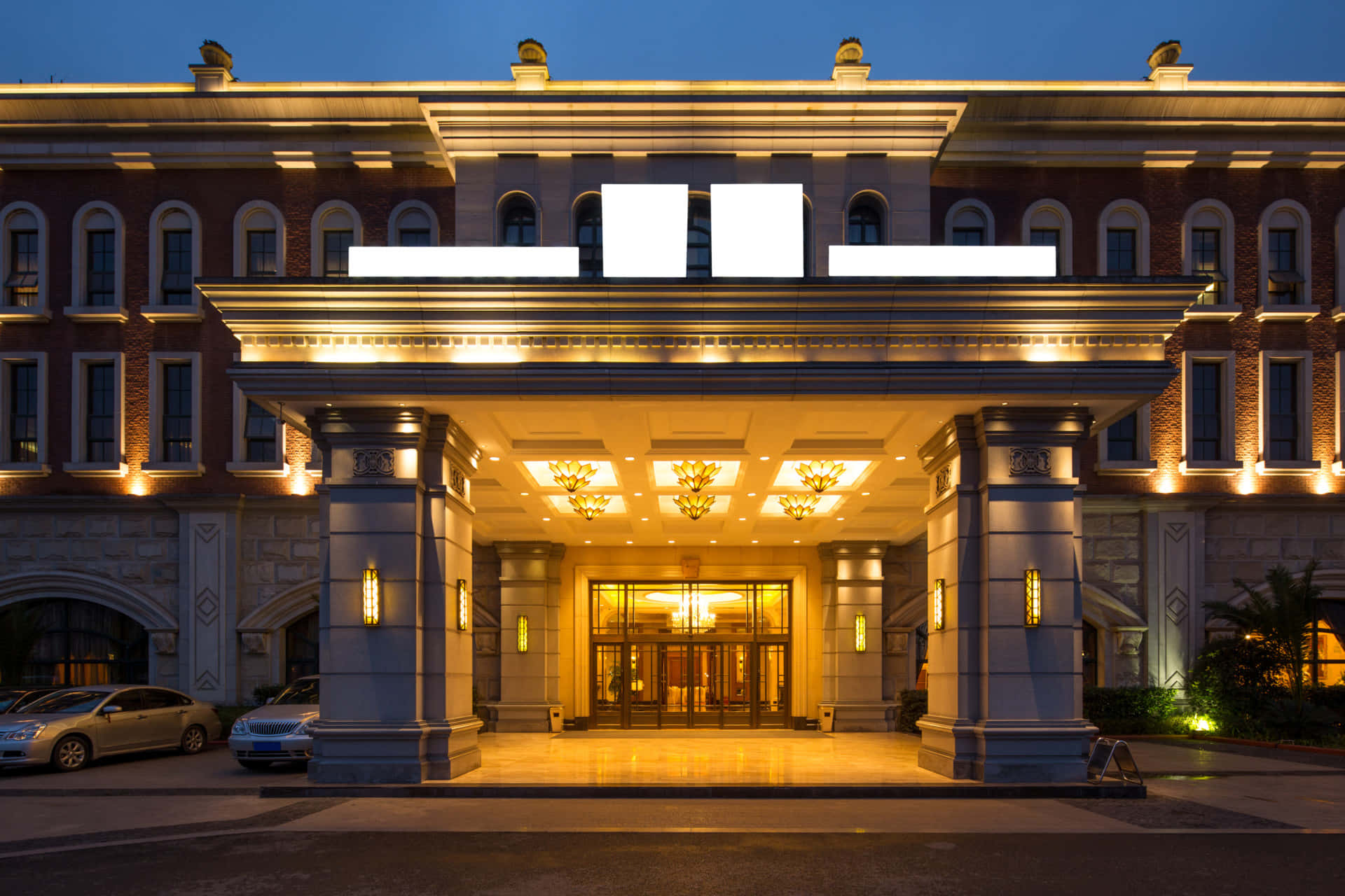 Hotel 桌布 壁紙 — 宏偉酒店入口在黃昏時亮起溫暖的金色燈光，映照在拋光石頭和停泊的汽車上。
