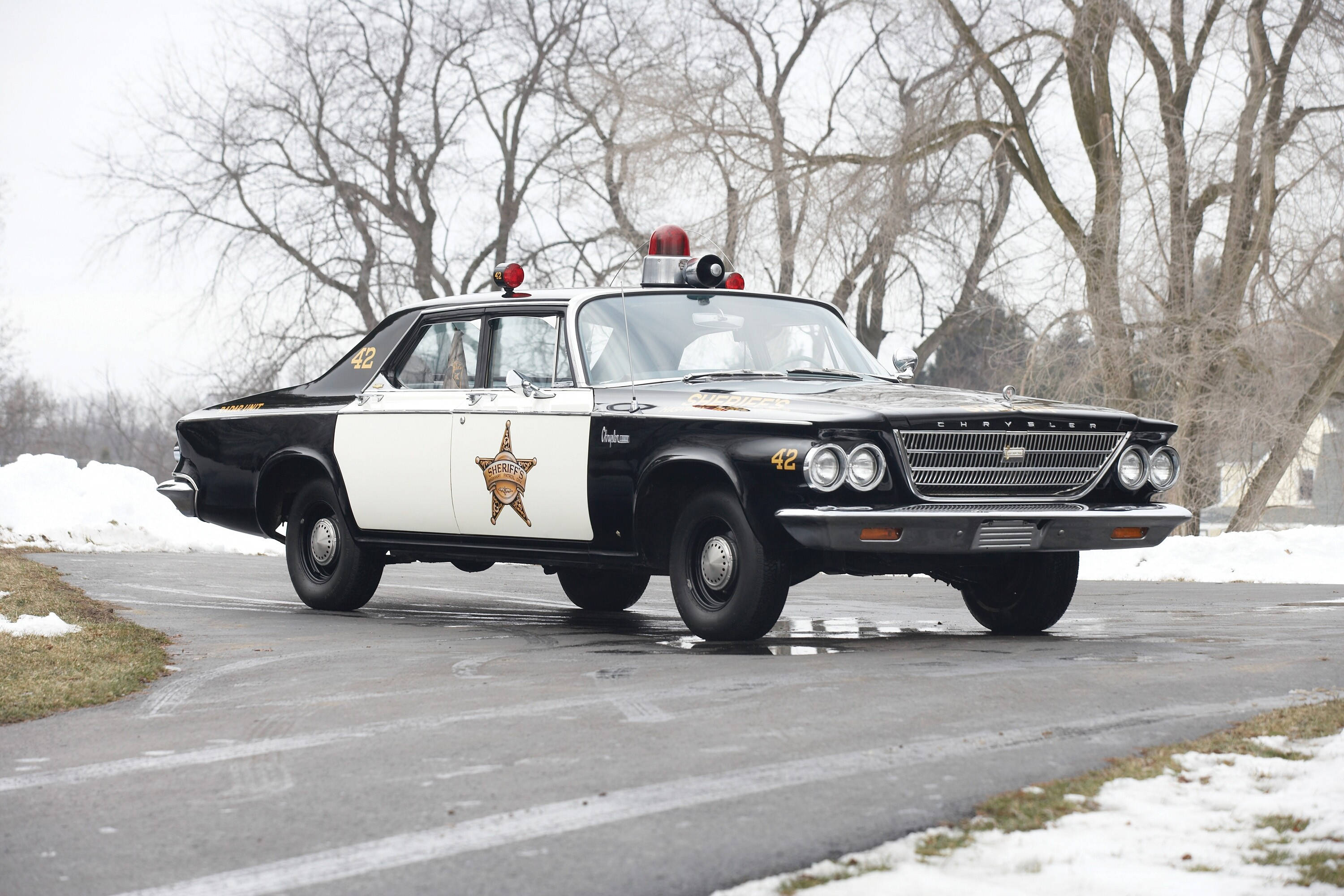 官方道奇警用巡邏車 壁紙 — 一輛帶有星形徽章的復古黑白警長車在積雪的道路上行駛，道路兩旁是光禿禿的樹木。