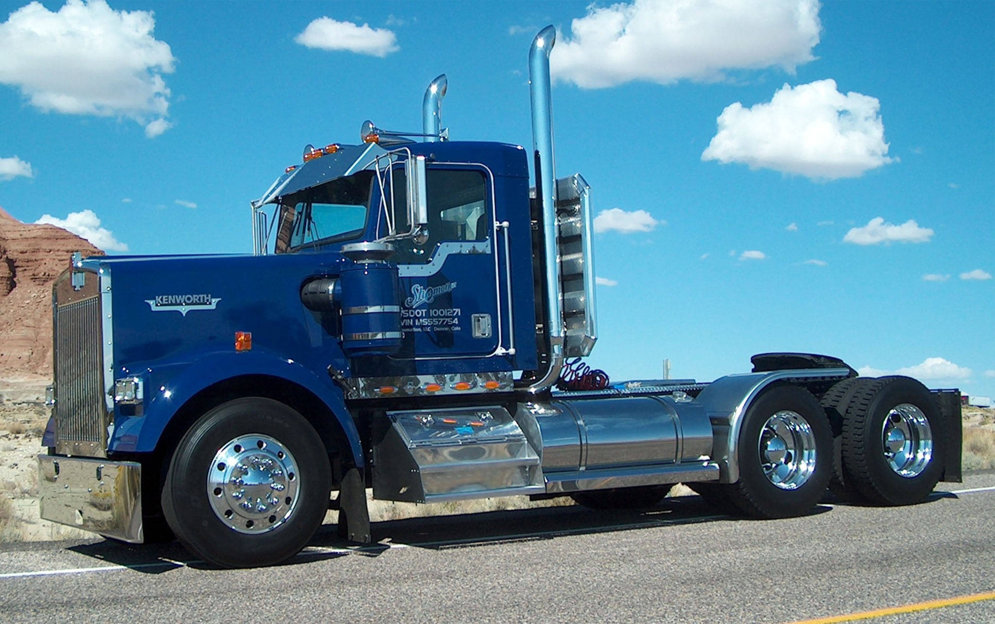 定制肯沃斯W900 壁紙 — 一輛閃亮的藍色 Kenworth 半掛卡車，帶有鍍鉻裝飾，停在沙漠公路上，天空湛藍，白雲朵朵。