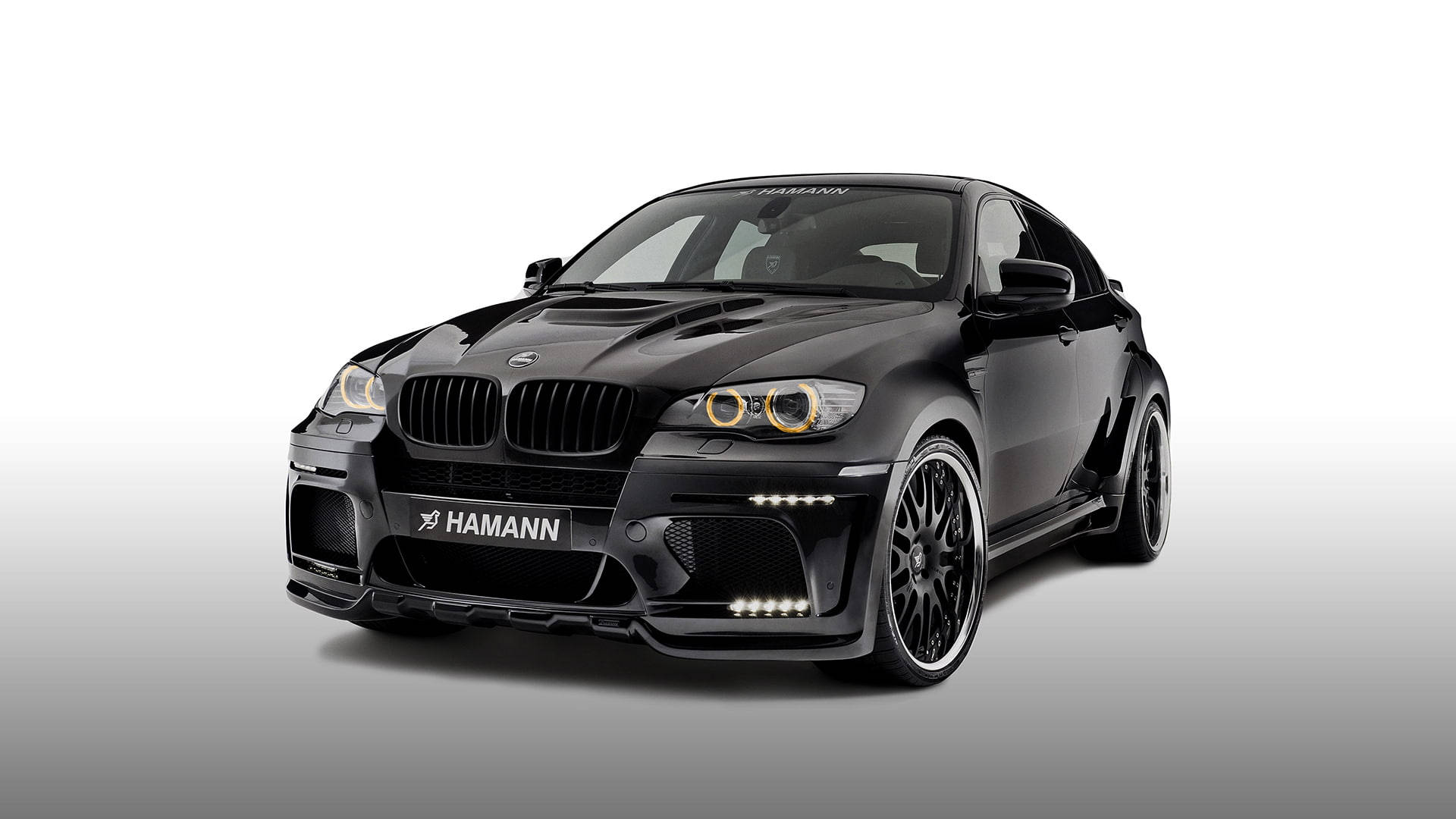客製化BMW X6 M 壁紙 — 一輛時尚的黑色 Hamann 改裝寶馬跑車，配備發光頭燈和定制輪圈，在漸變工作室背景前擺拍。