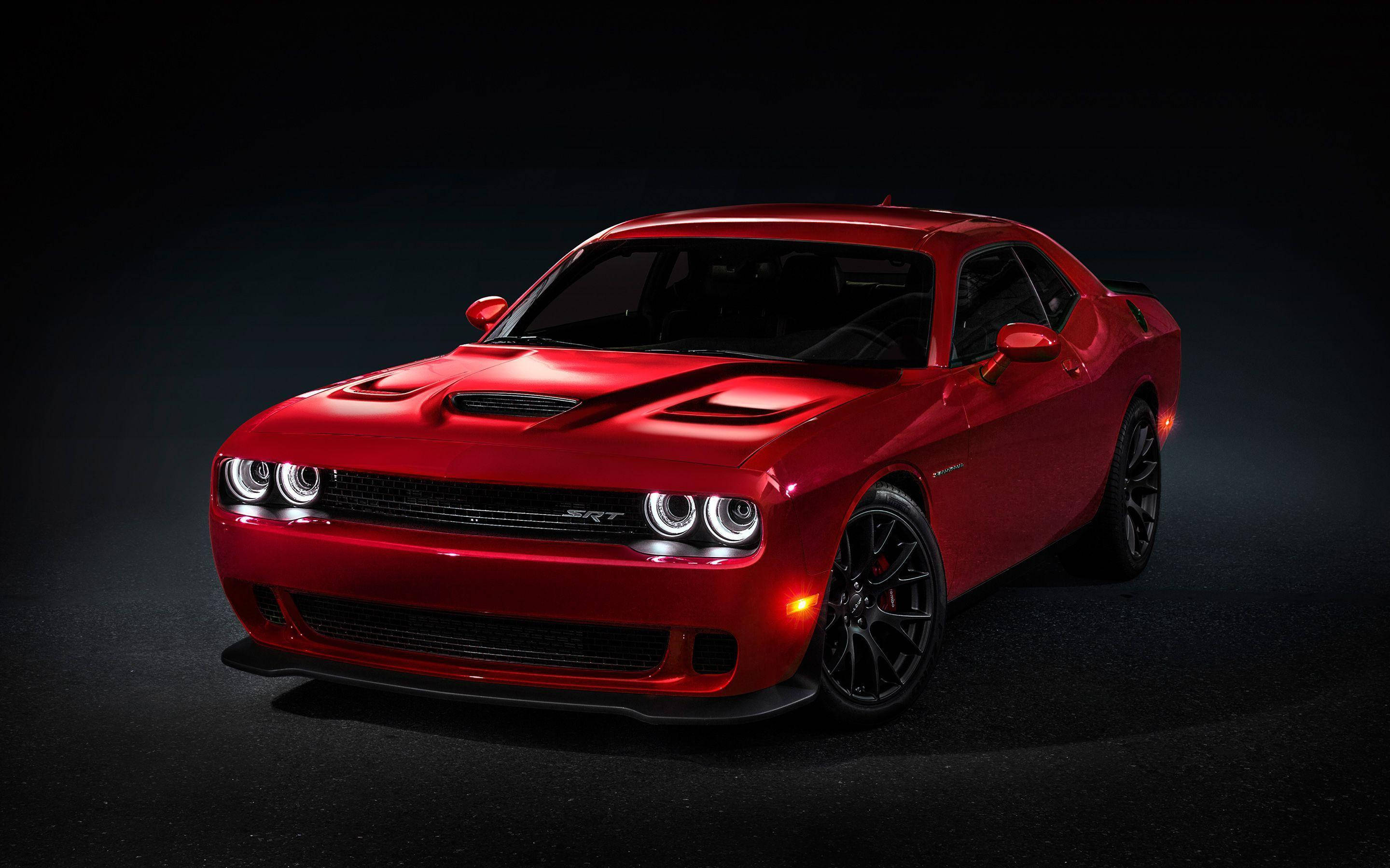 客製化道奇挑戰者Unleashed 壁紙 — 一輛光澤的紅色Dodge Challenger SRT，帶有發光的頭燈和黑色輪圈，在黑暗的攝影棚背景下戲劇性地照亮。
