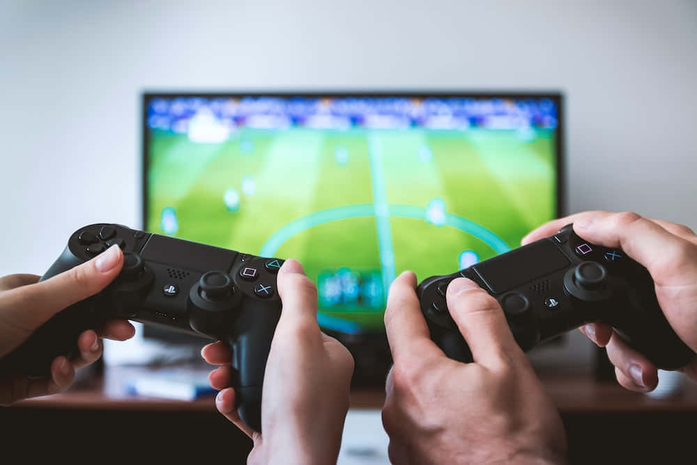 宣佈 Sony PlayStation，連結全球玩家 壁紙 — 兩隻手緊握黑色遊戲控制器，專注於電視屏幕上顯示的一場充滿活力的足球比賽。