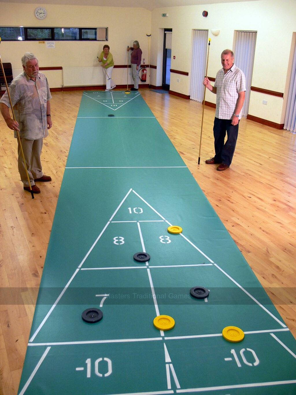 室內 Shuffleboard 遊戲進行中 壁紙 — 四位長者在明亮的木地板大廳中，在標有編號區域的綠色球場上玩沙狐球。