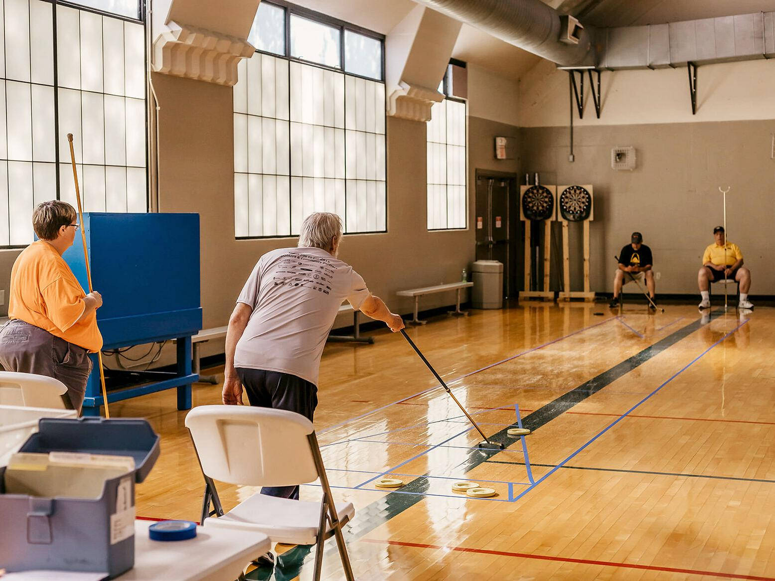 室內地板 Shuffleboard 遊戲的熱烈時刻 壁紙 — 兩位長者在拋光木地板上玩沙狐球，其他人在附近觀看。