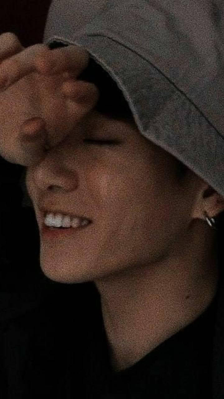 害羞 Jungkook 美學 壁紙 — 一個戴著灰色漁夫帽嘅人喺昏暗燈光下用一隻手遮住對眼，露出溫暖嘅笑容。