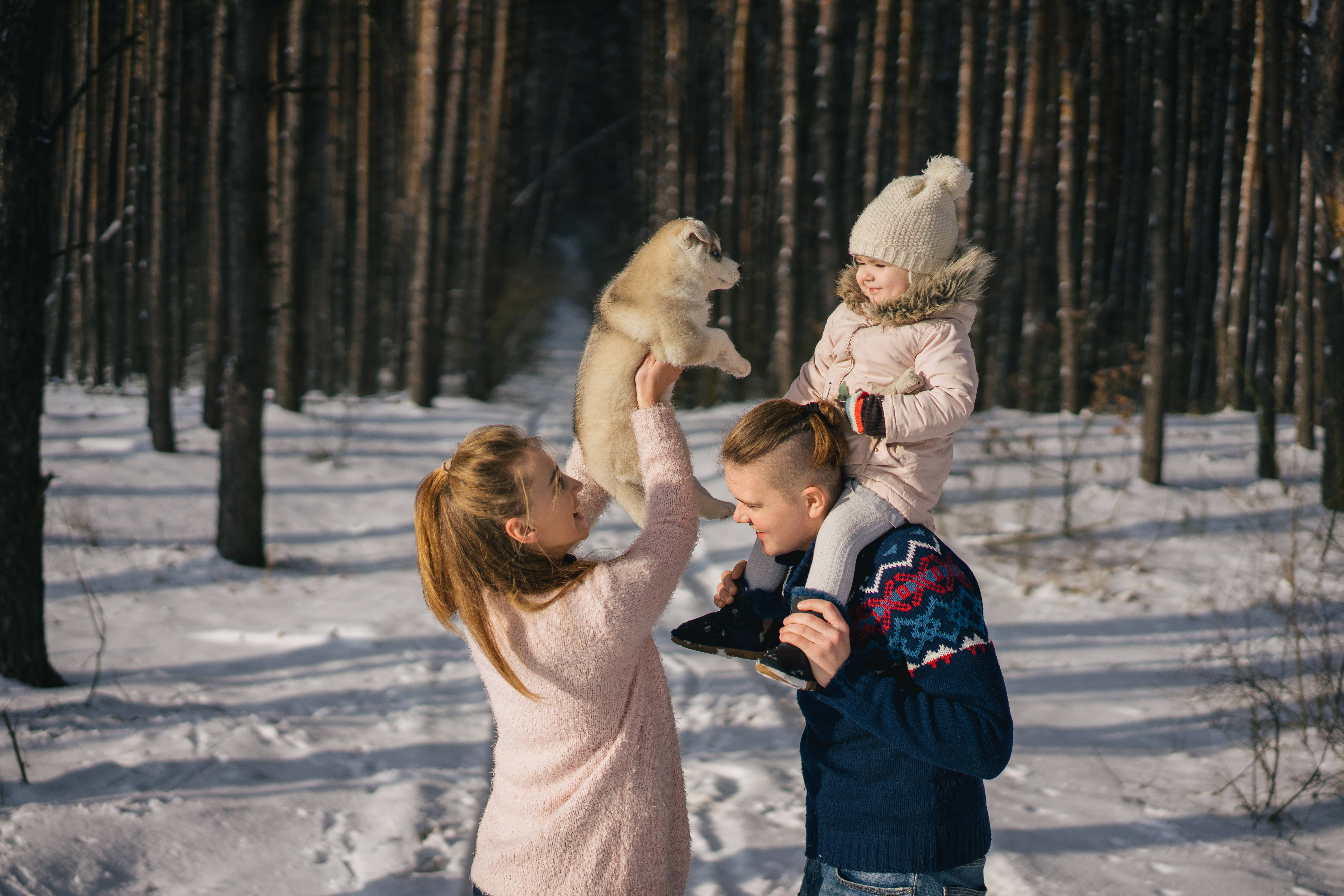 家人冬季森林之旅 壁紙 — 在雪地森林中，母親抱著哈士奇幼犬，父親背著孩子，一個歡樂的冬季家庭時刻。
