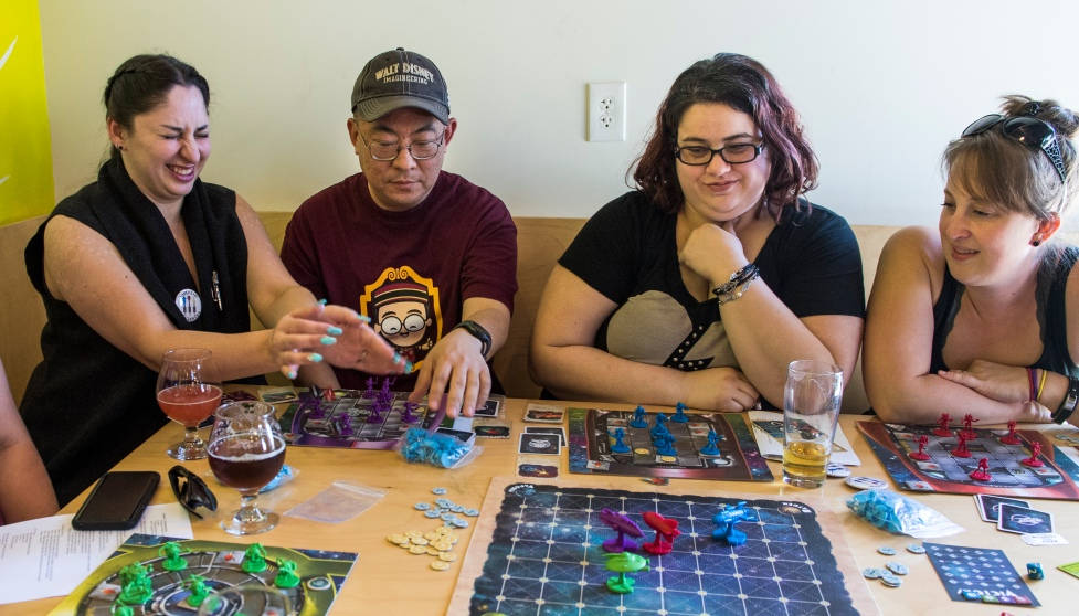 四人在桌旁玩棋盤遊戲，邊喝飲料邊笑。