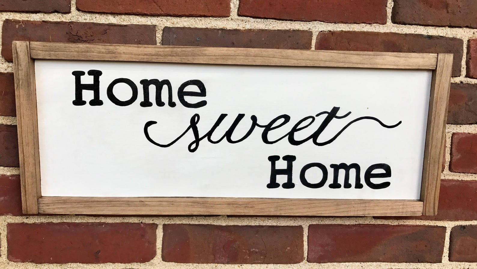 家園甜蜜歡迎板 壁紙 — 一個質樸的木牌，上面用黑色草書寫著「Home sweet Home」，掛在紅磚牆上。