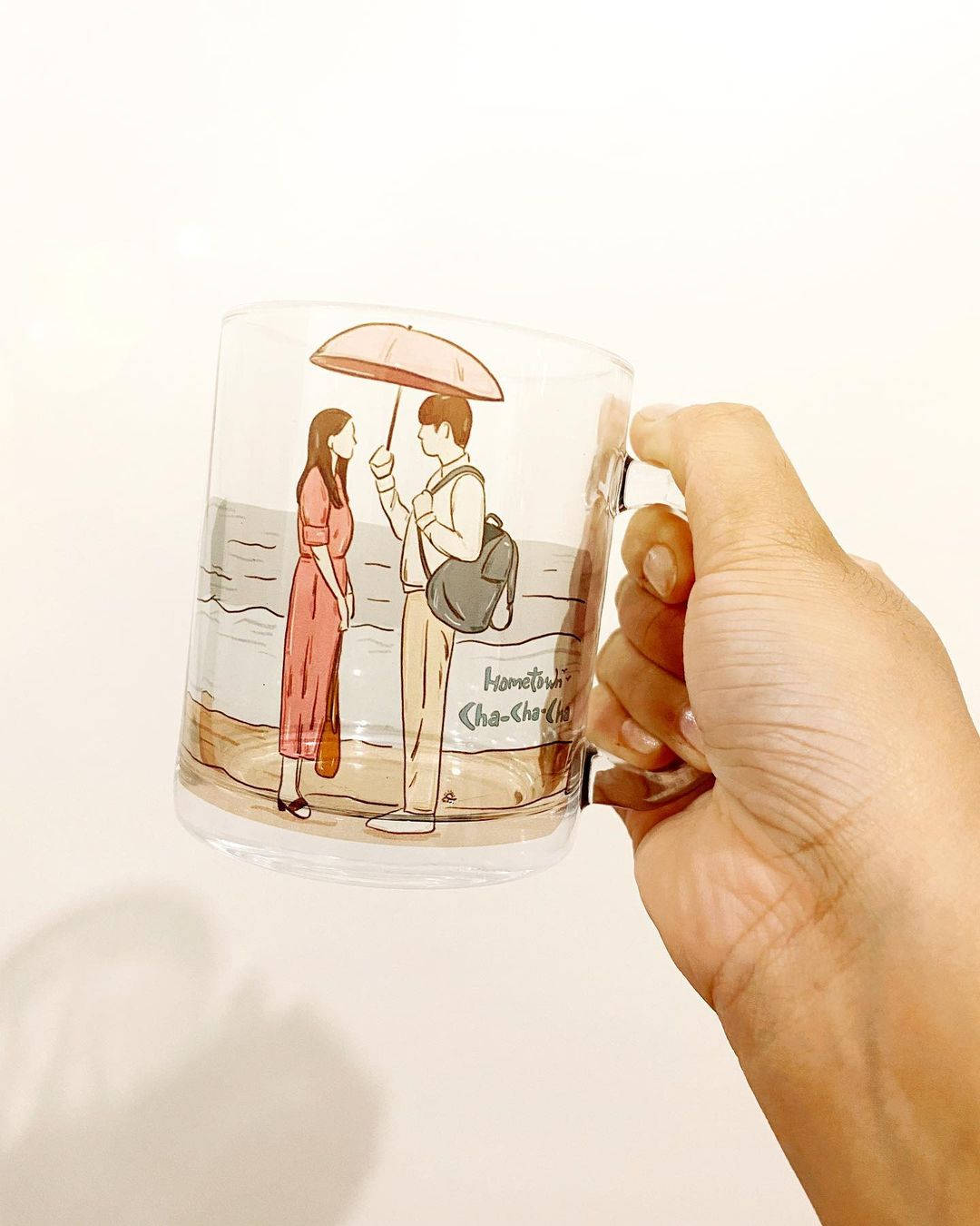 《家在首爾》周邊商品 壁紙 — 一隻手拿著一個透明馬克杯，上面有插畫情侶在海灘上撐傘，標有'Hometown Cha-Cha-Cha'。