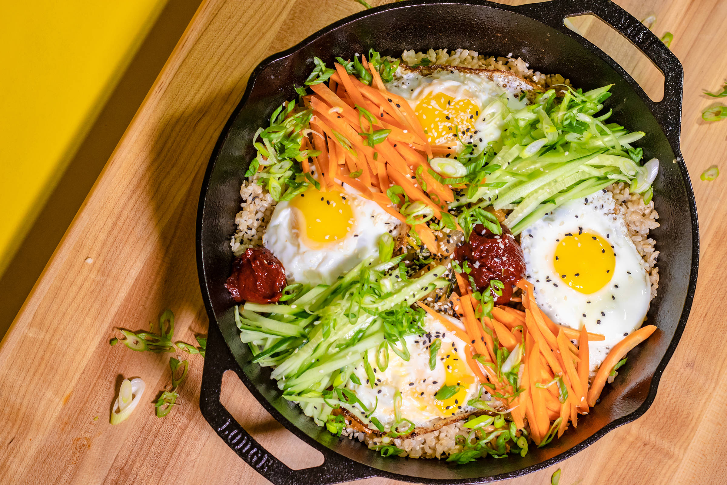 家庭份量韓式拌飯 壁紙 — 一個充滿活力的韓國石鍋拌飯，放在黑色鑄鐵煎鍋中，上面放有太陽蛋、刨絲紅蘿蔔、青瓜和韓式辣醬，置於木板上。