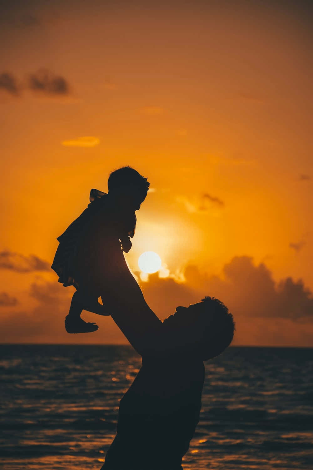 家庭共享愛在父親節 壁紙 — 父親在海洋上將孩子高高舉起，映襯著充滿活力的橙色夕陽的剪影。