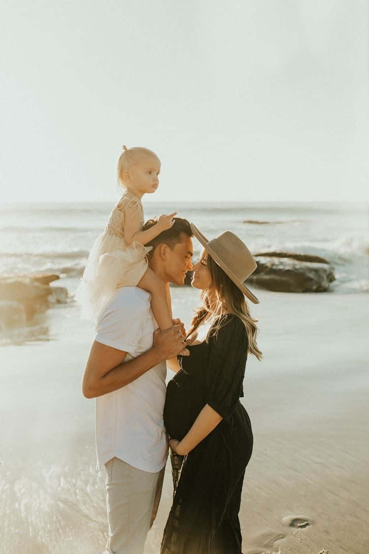 一對懷孕的夫婦和他們年幼的孩子在陽光明媚的海灘上擁抱。