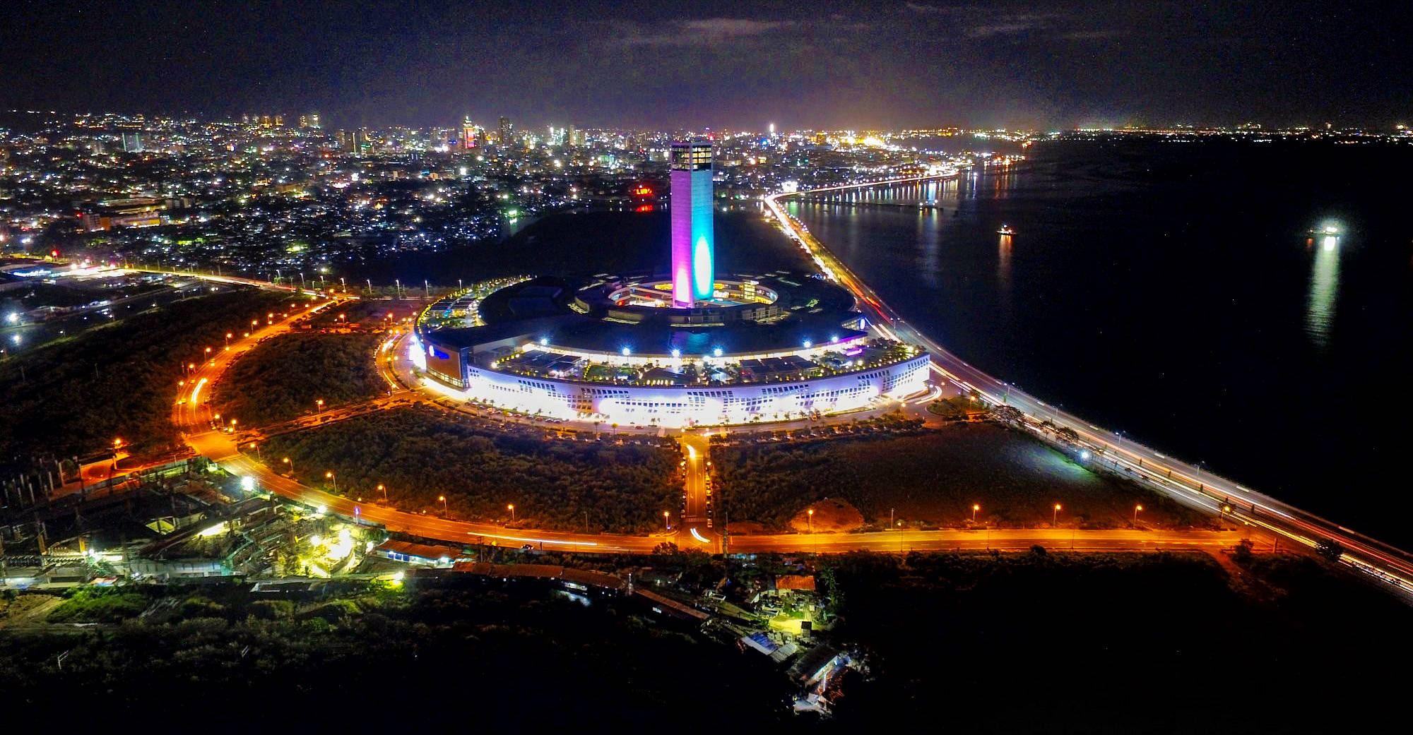 宿霧市SM海濱夜景 壁紙 — 夜晚鳥瞰一座明亮照明的圓形建築，旁邊有一座色彩繽紛的塔樓，毗鄰海濱城市景觀。