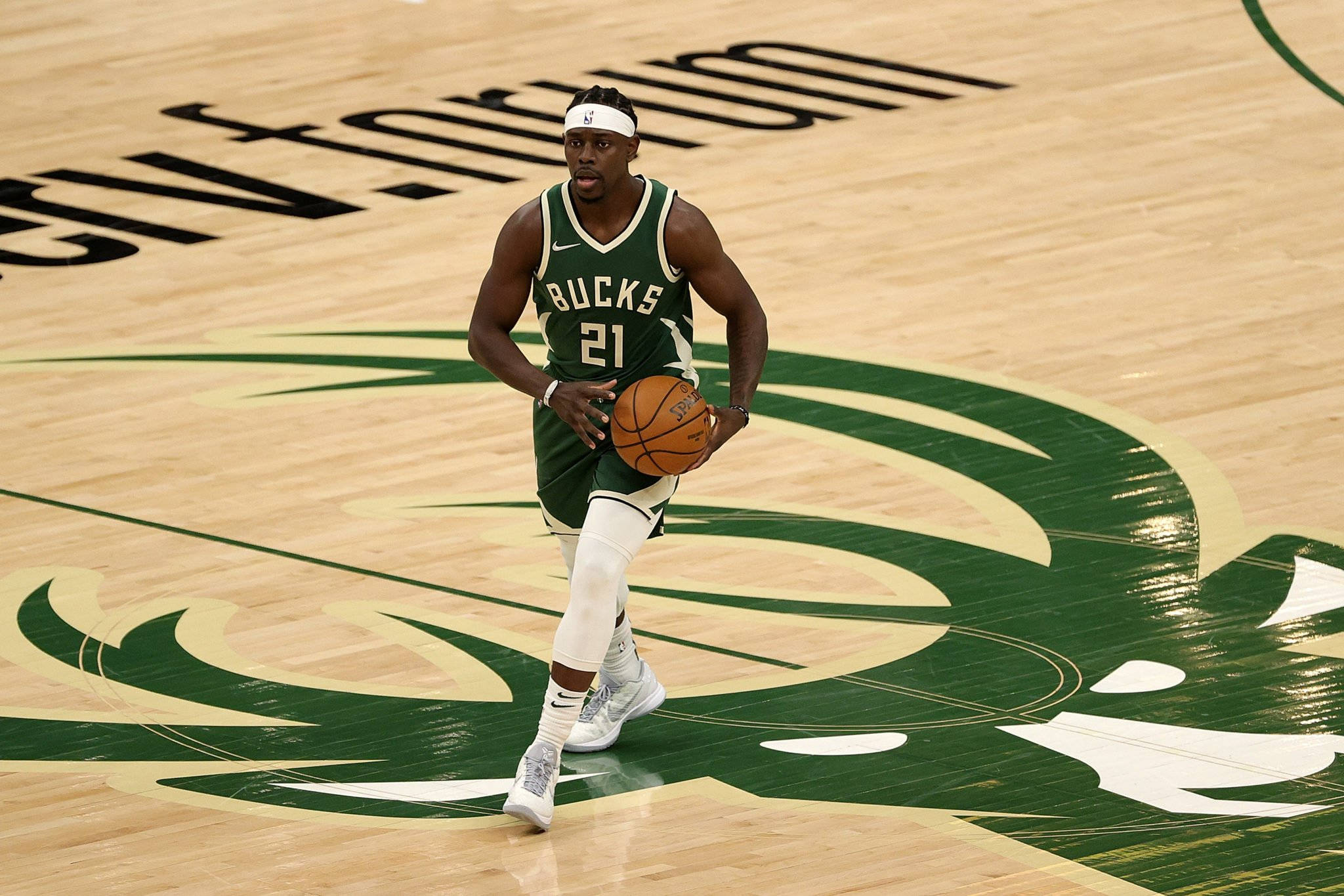 密爾沃基雄鹿隊Jrue Holiday 壁紙 — 一位身穿綠色球衣的密爾沃基公鹿籃球運動員在帶有球隊標誌的拋光木地板上運球。