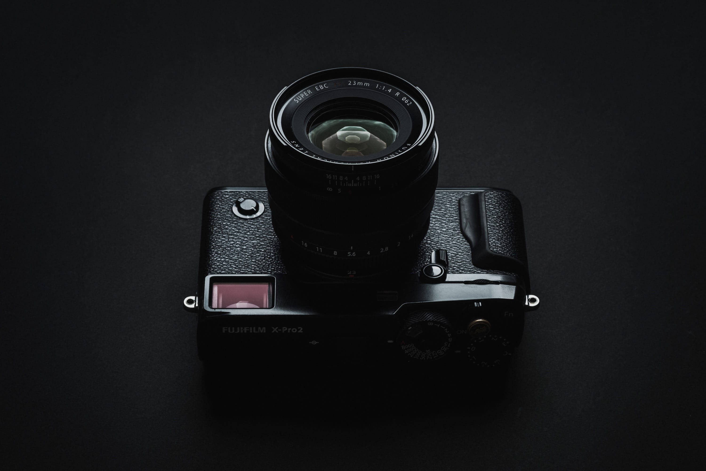 富士X-Pro2相機 壁紙 — 一部黑色Fujifilm X-Pro2相機配備23mm鏡頭，置於深色背景中央，從上方打光以突顯其紋理機身和金屬轉盤。