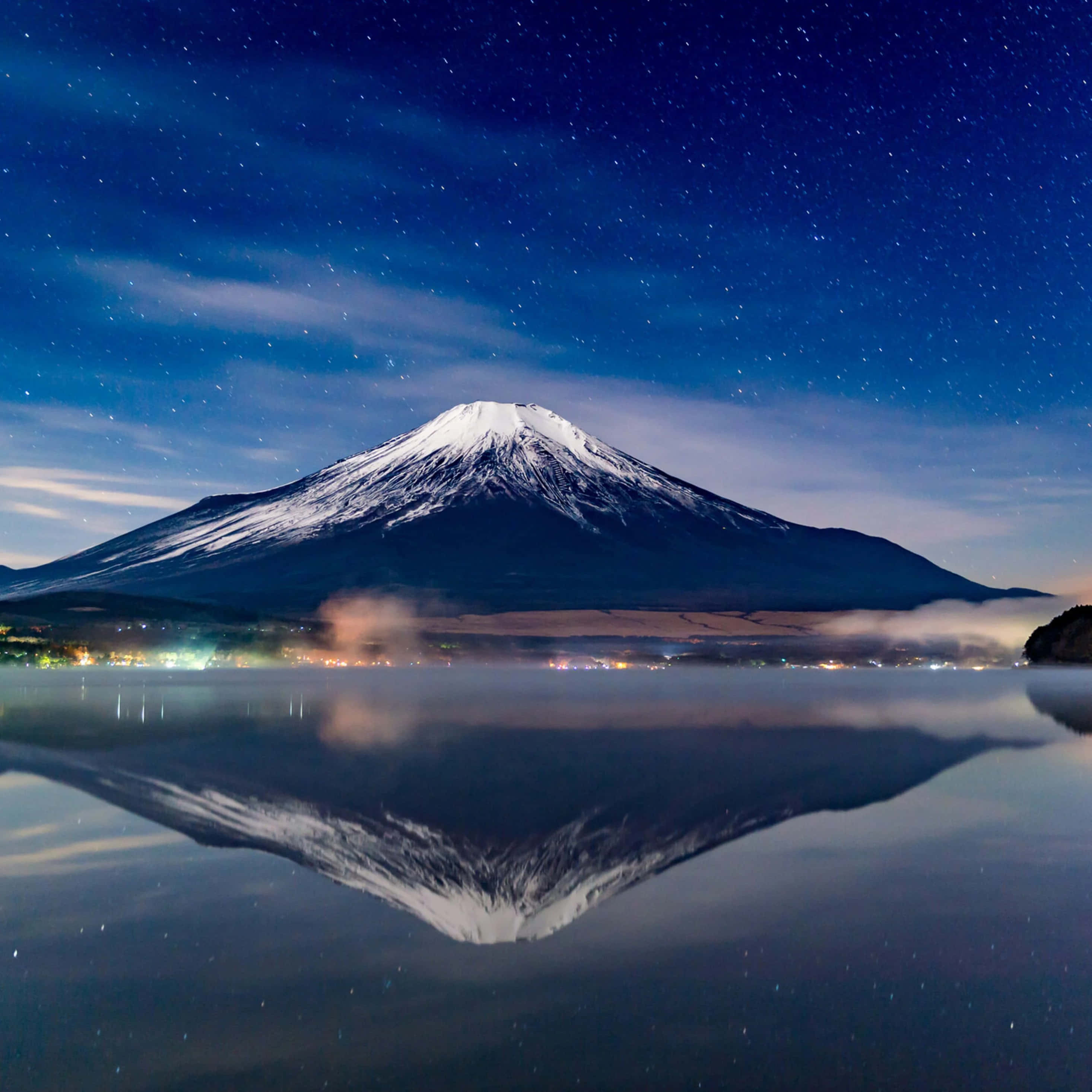富士山，日本的象徵性山峰 壁紙 — 白雪覆蓋的富士山在平靜的湖面上完美倒映，下方是繁星點點的夜空和微弱的城市燈光。