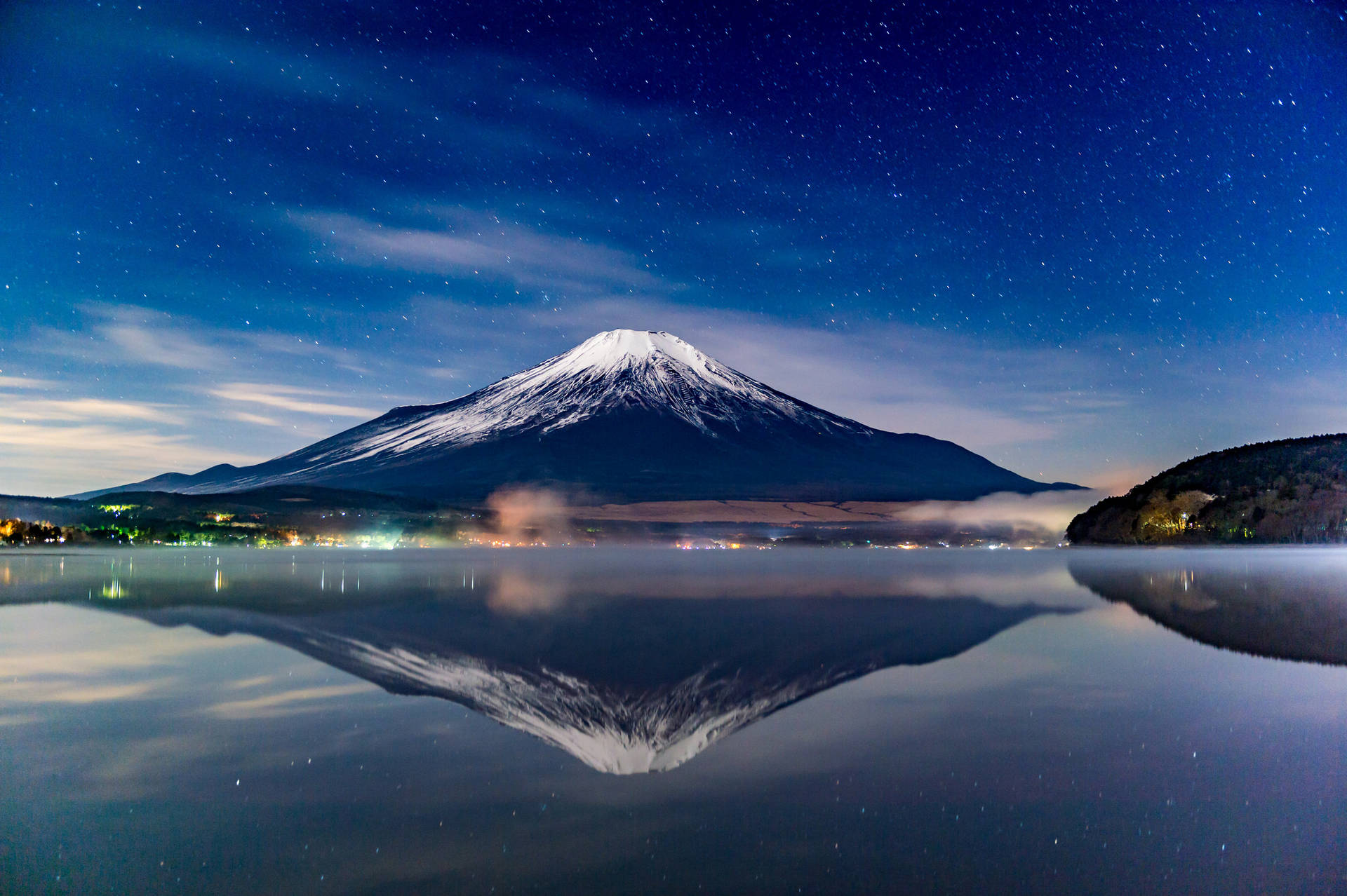 富士山在星空下平靜的湖面上倒映，遠方燈光點點。