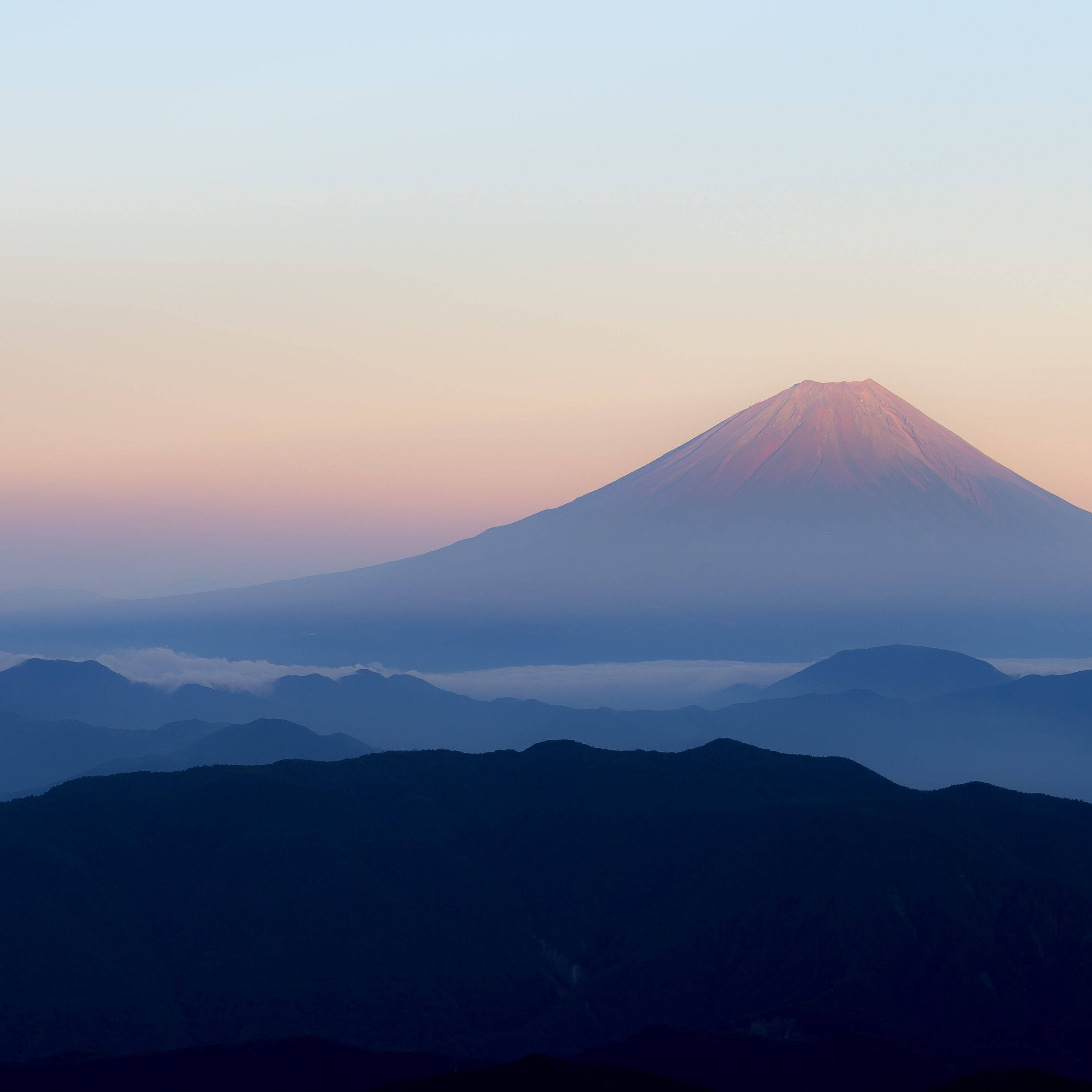 HD Mountain 桌布 壁紙 — 富士山在黎明時分雄偉地升起，柔和的粉紅色和藍色色調在層疊的山巒剪影上空繪畫天空。