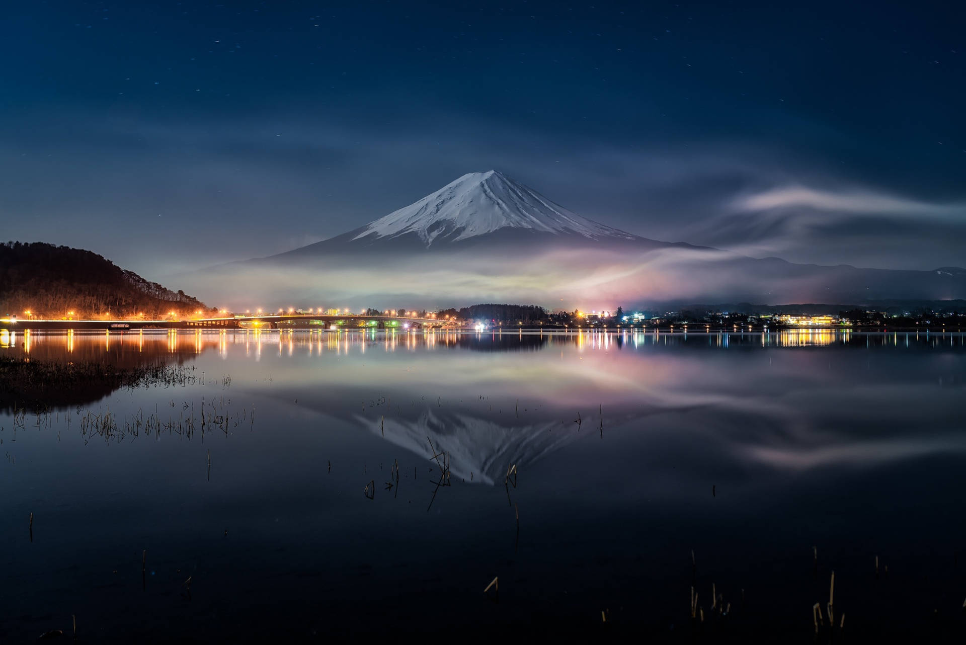 富士山在星空下的平靜湖面倒映，遠處有城市燈光。