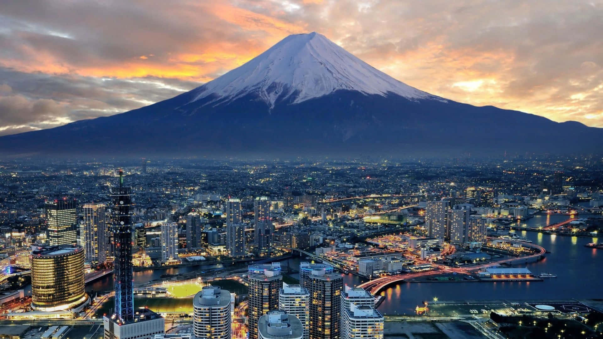 富士山與東京夜景 壁紙 — 白雪覆蓋的富士山在日落時分聳立在發光的城市景觀之上，下方有溫暖的橙色雲彩和閃爍的燈光。