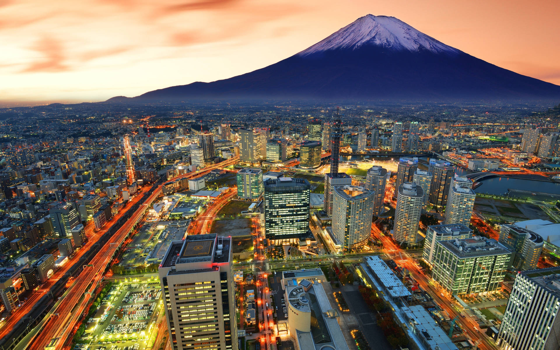 富士山與橫濱市景 壁紙 — 白雪覆蓋的富士山在日落時分聳立於廣闊的城市景觀之上，伴隨著閃爍的燈光和流動的車流。