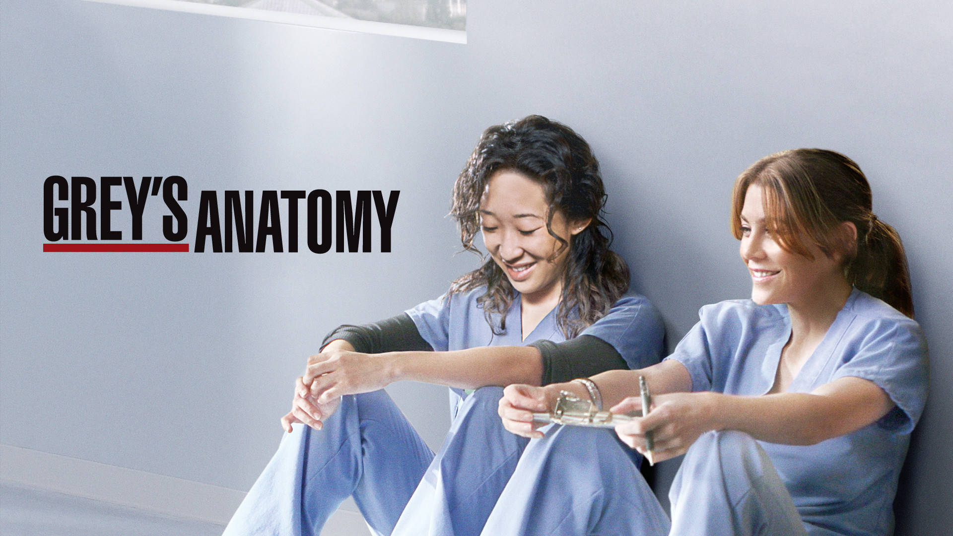 兩位穿著藍色手術服的女人微笑著坐在牆前，牆上有「Grey's Anatomy」文字。