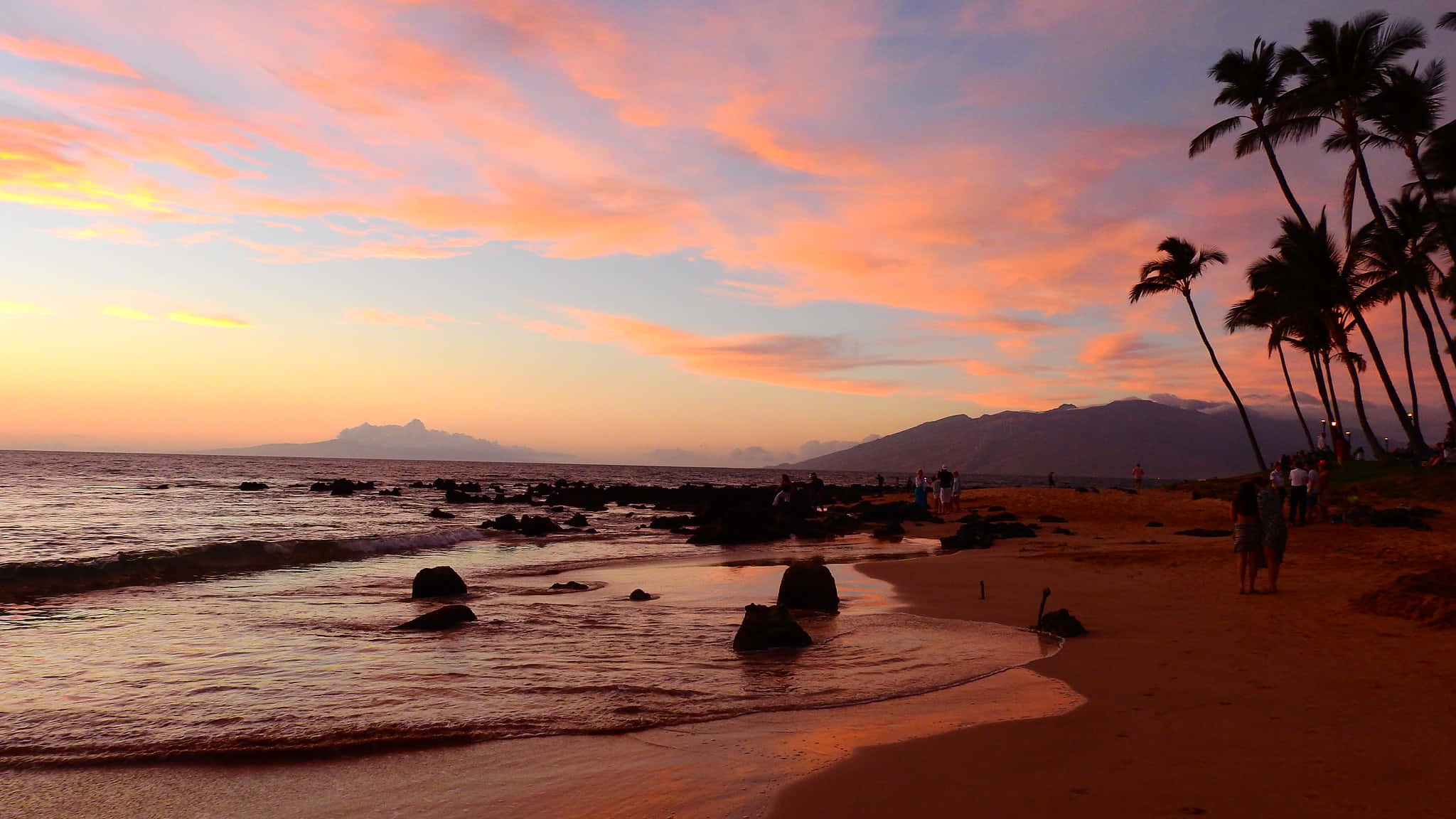 寧靜夏威夷夕陽風景 壁紙 — 充滿活力的日落用火熱的橙色和粉紅色調描繪天空，映照在一個有棕櫚樹和遠山的熱帶海灘上。