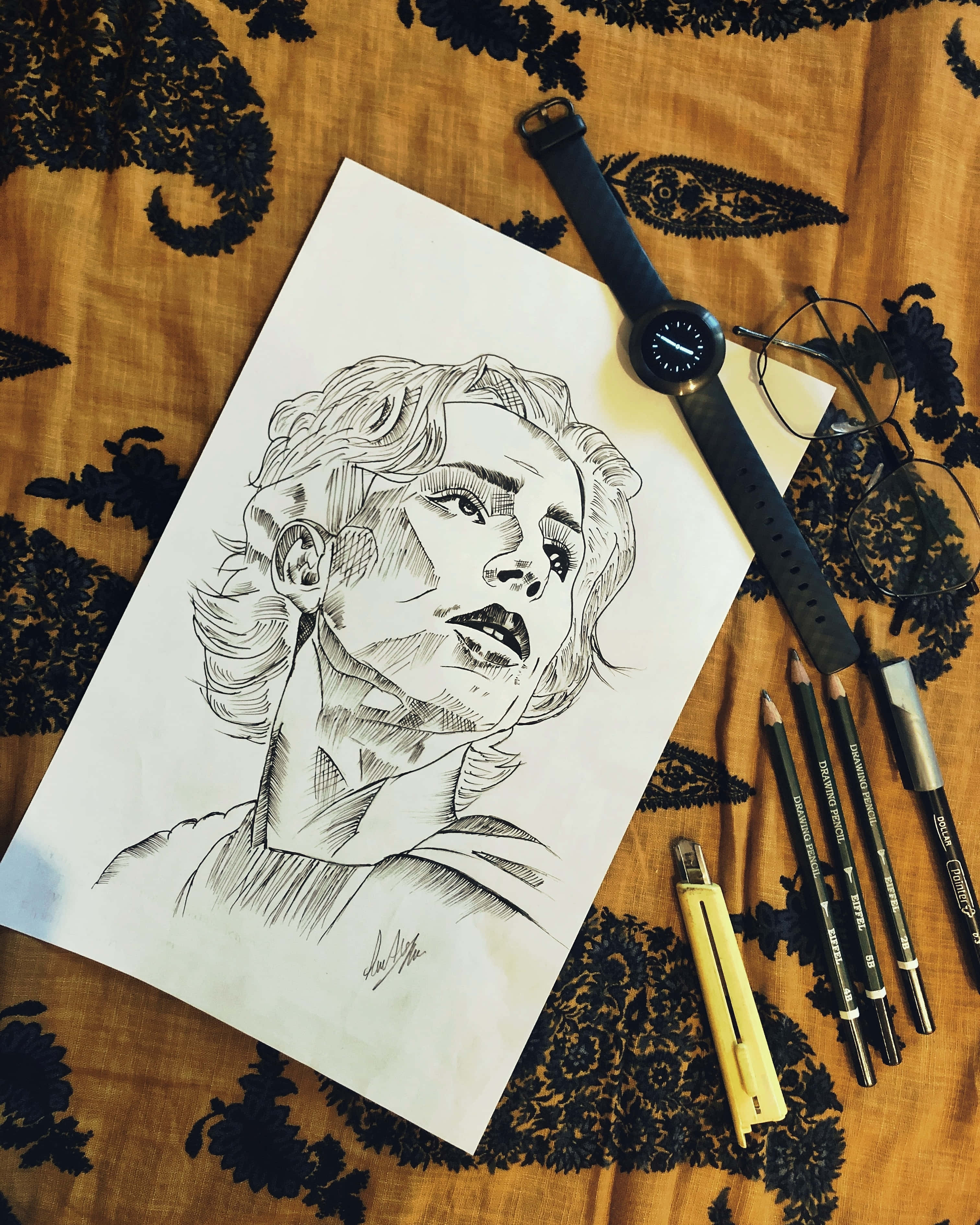 寧靜山景 壁紙 — 一幅詳細的黑白鉛筆素描，描繪一個人的臉部，放在有圖案的布料上，旁邊有繪畫工具和智能手錶。